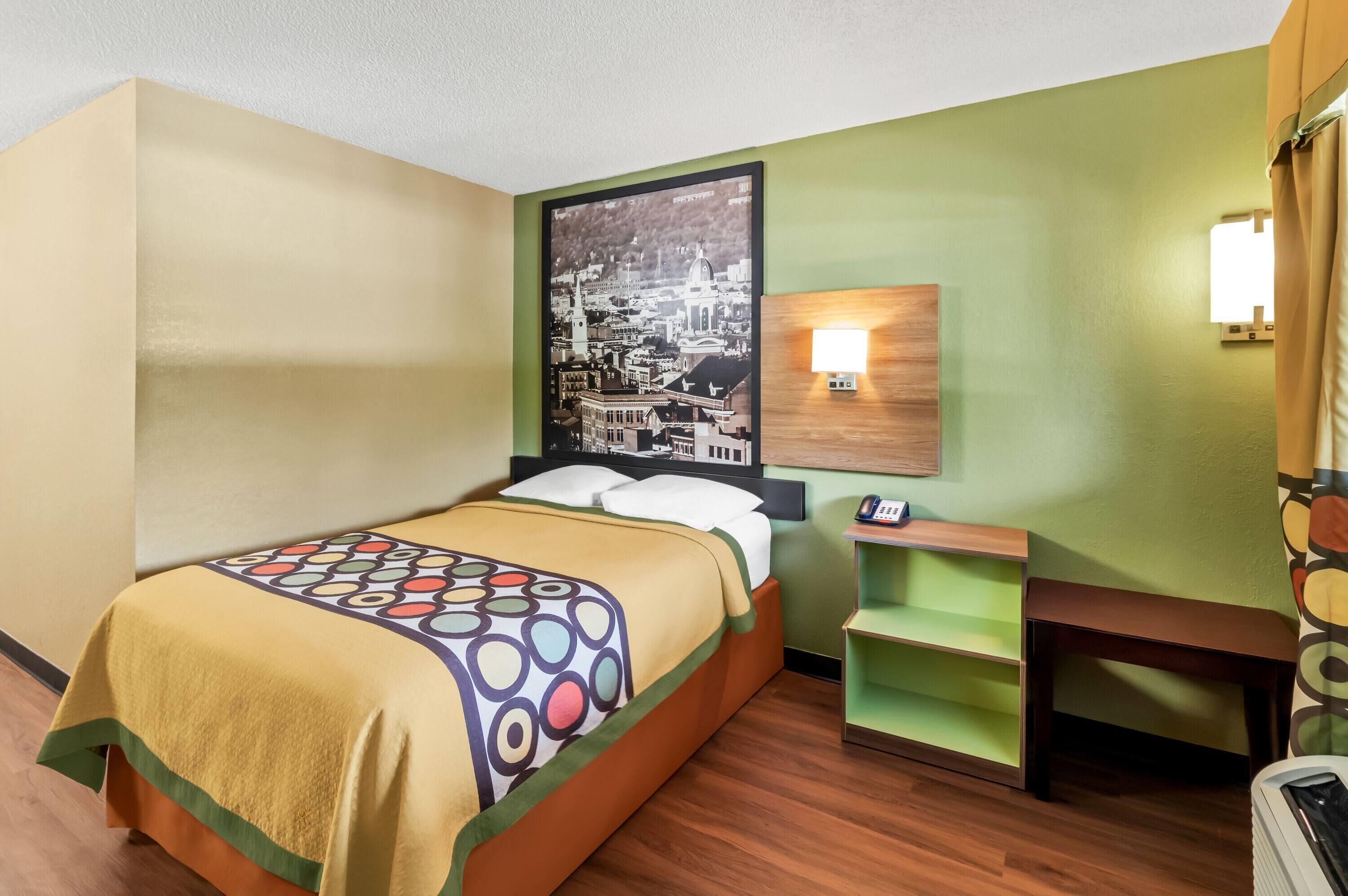 A hotel room with a bed, a wall-mounted lamp, a small wooden table, and a green shelf.
