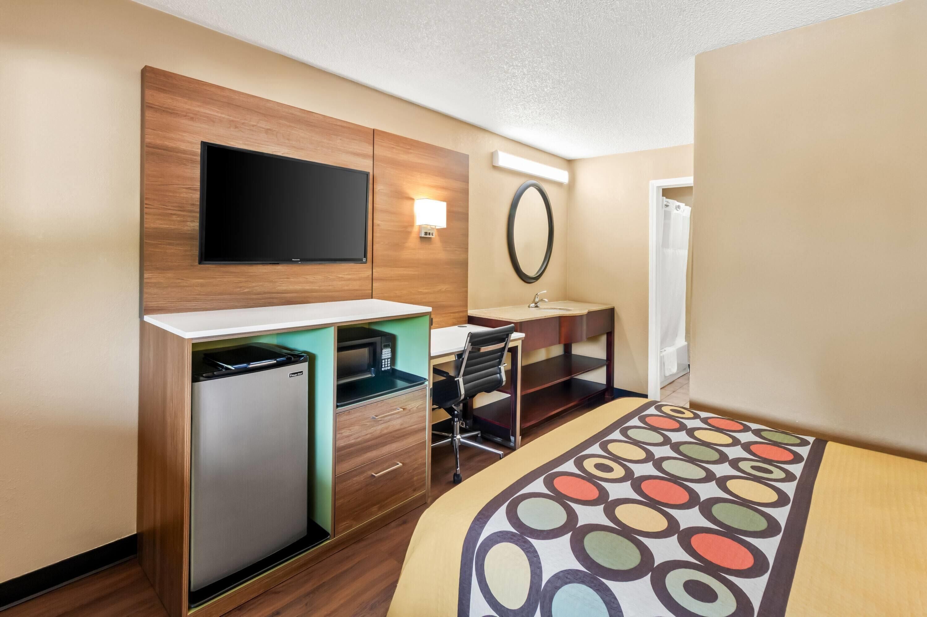 Habitación de hotel compacta con cama, escritorio con computadora, refrigerador pequeño, televisor y baño con ducha.