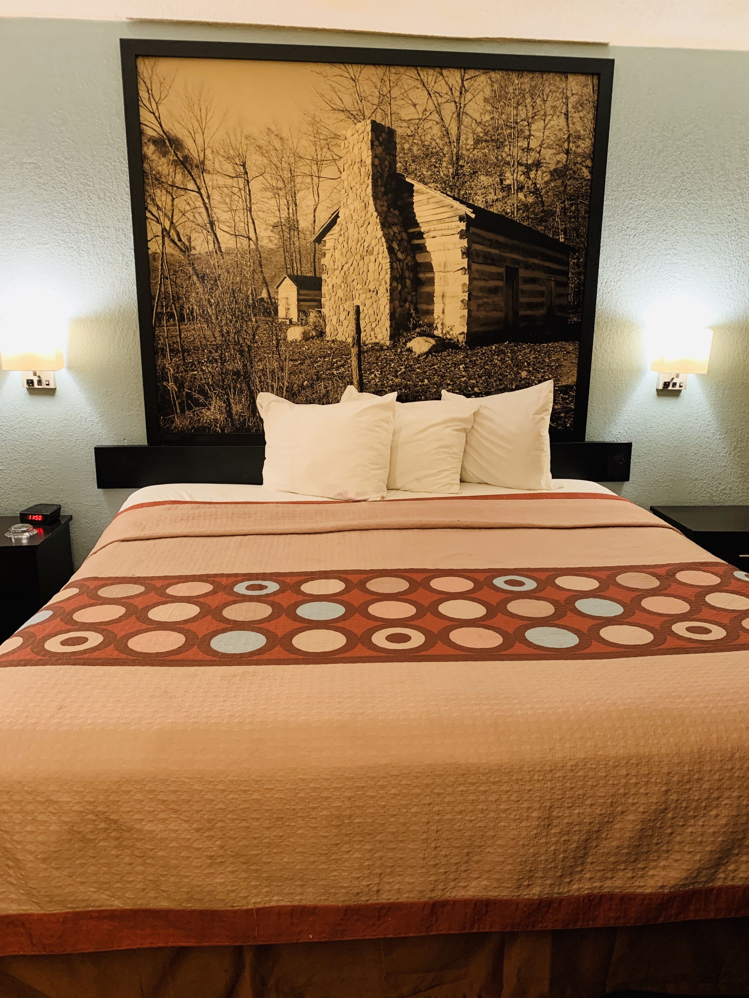 A hotel room with a large bed, a headboard with a framed picture, two bedside lamps, and a clock on the nightstand.
