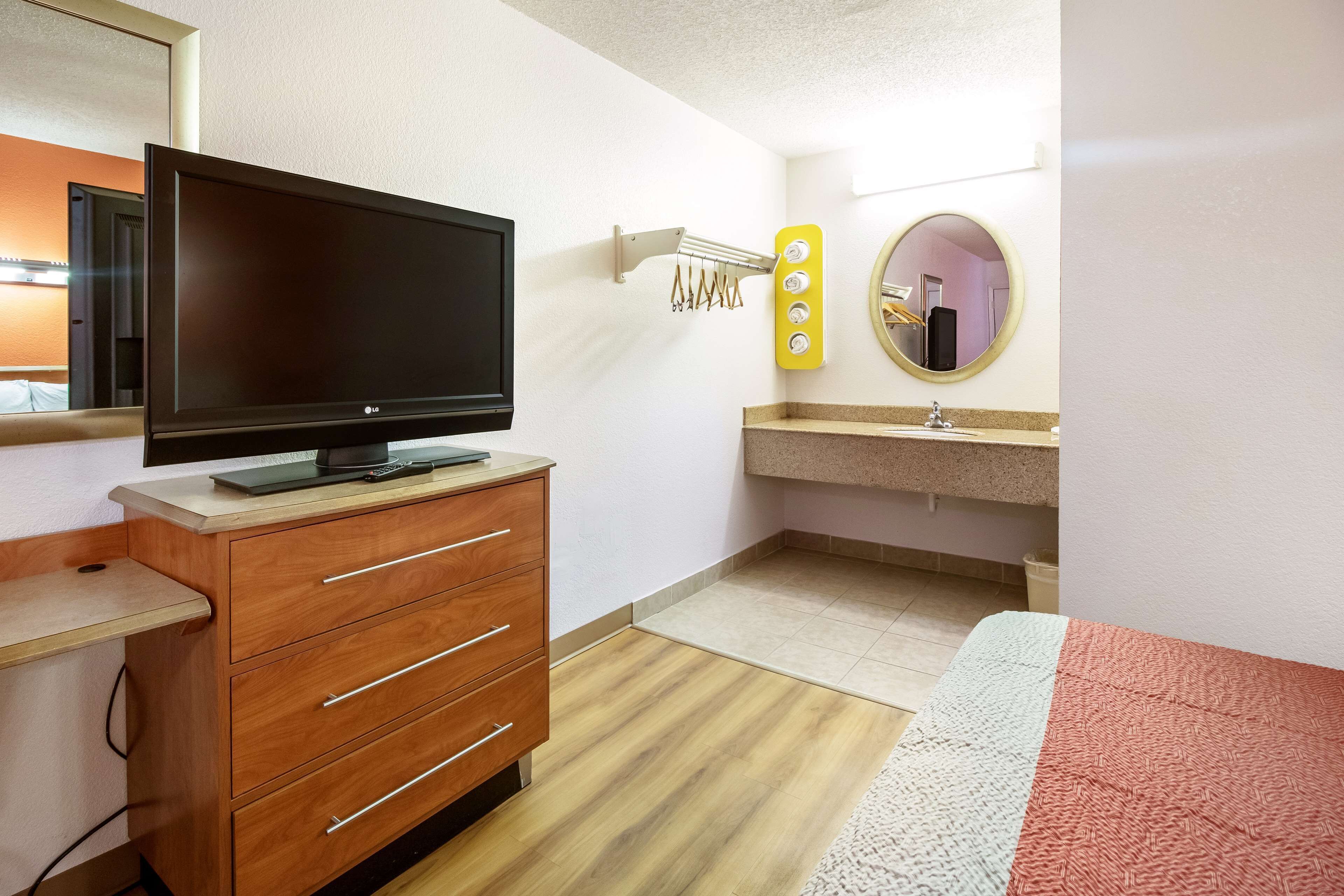 Habitación de hotel con televisor de pantalla plana sobre un tocador de madera, un espejo con luces, un lavamanos y una cama con una colcha roja.
