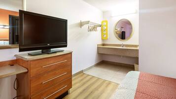 Ein Hotelzimmer mit Flachbildfernseher auf einer Holzkommode, einem Spiegel mit Beleuchtung, einem Waschbecken und einem Bett mit roter Decke.