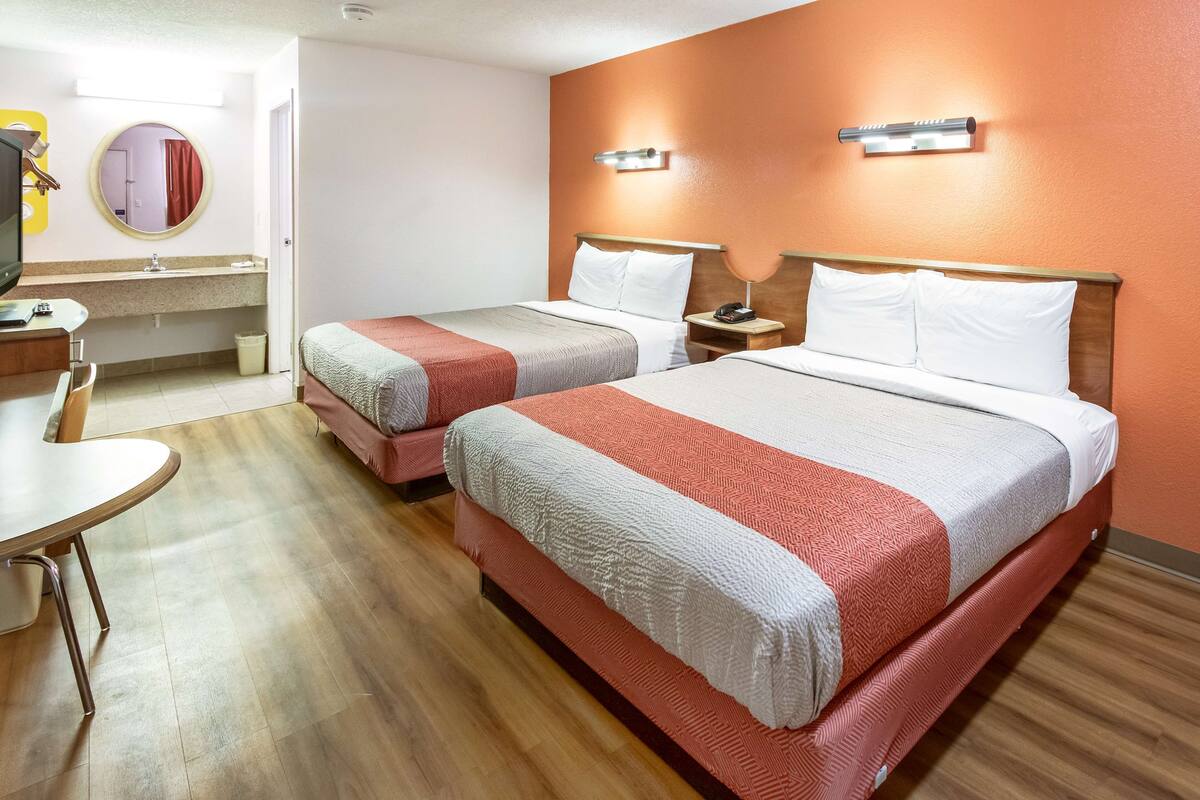 A hotel room with two beds, a desk, a chair, a mirror, and a bathroom.