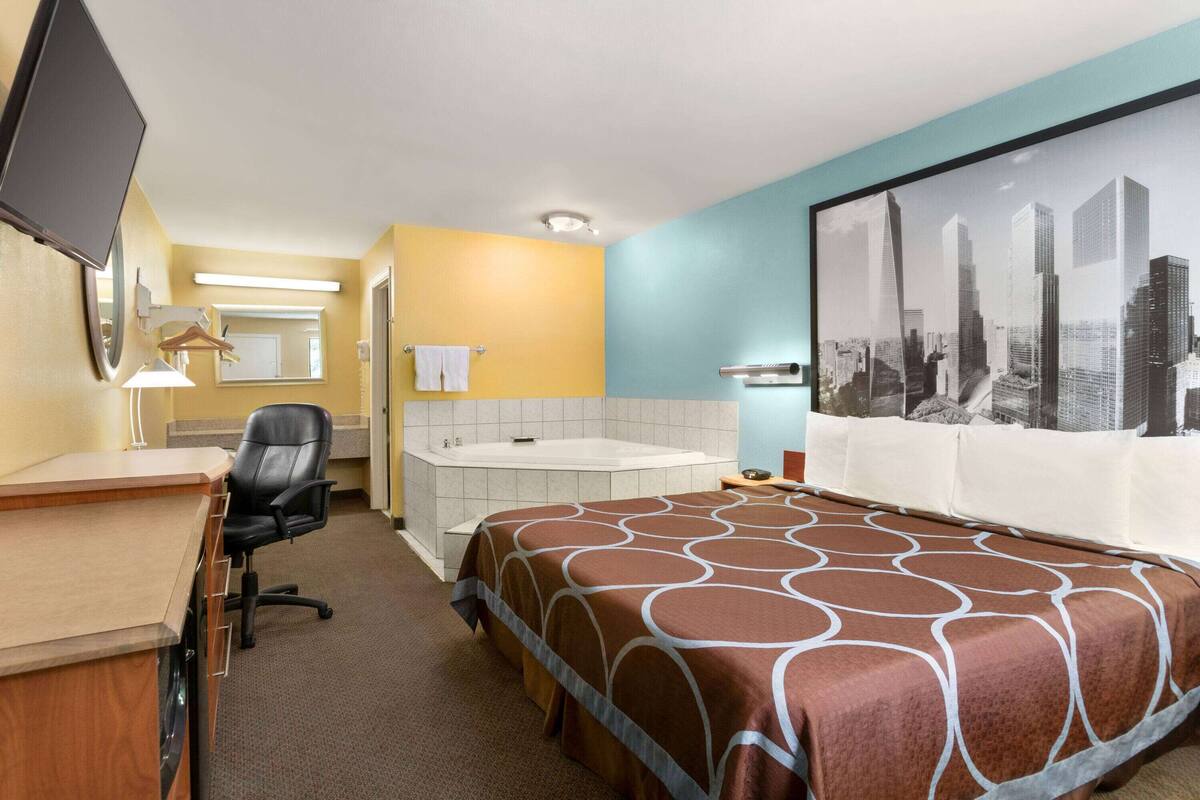 A hotel room with a bed, a desk, a chair, a bathtub, and a large cityscape painting on the wall.