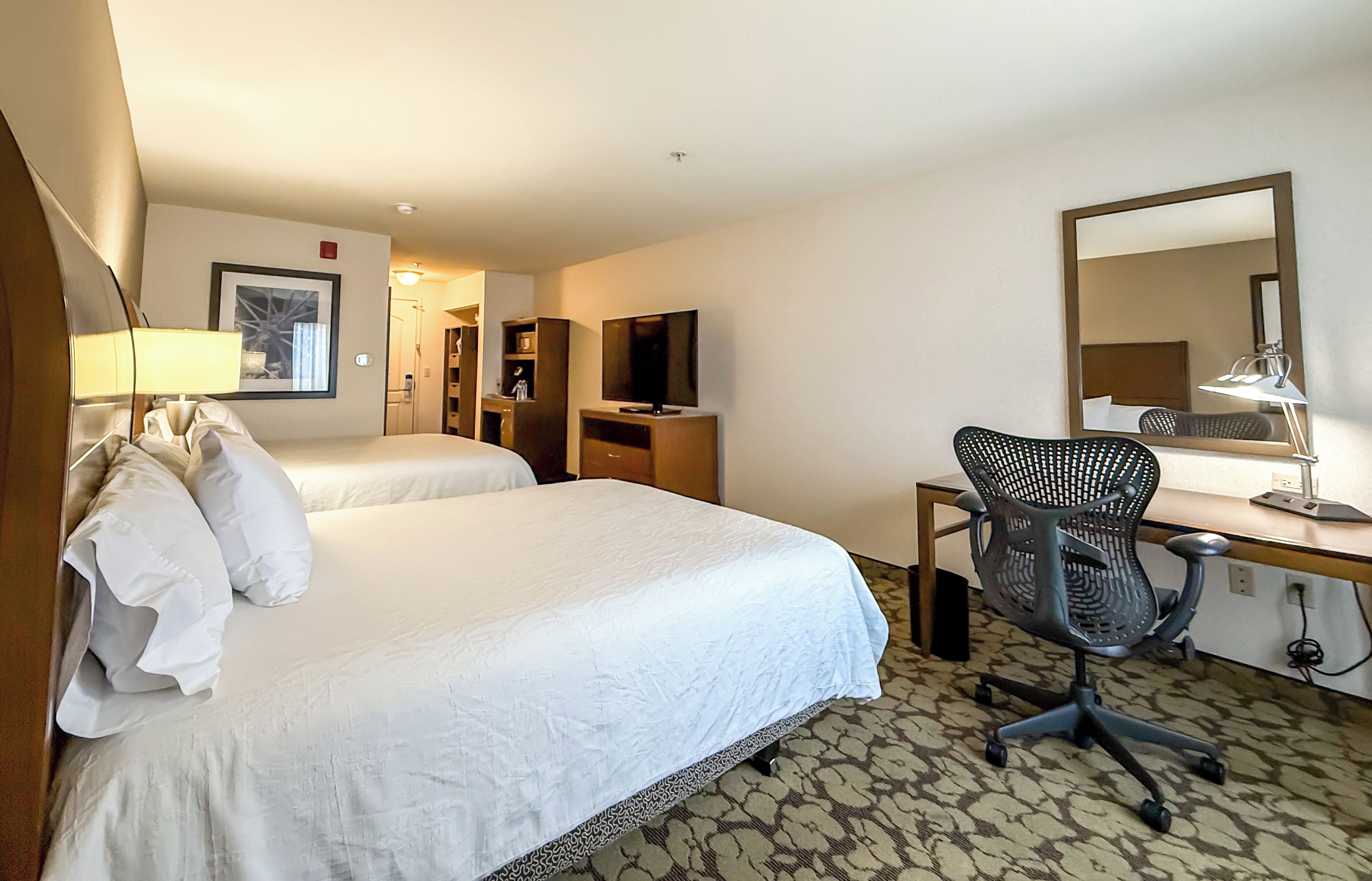 A hotel room with two beds, a desk with a chair, a TV, and a mirror.