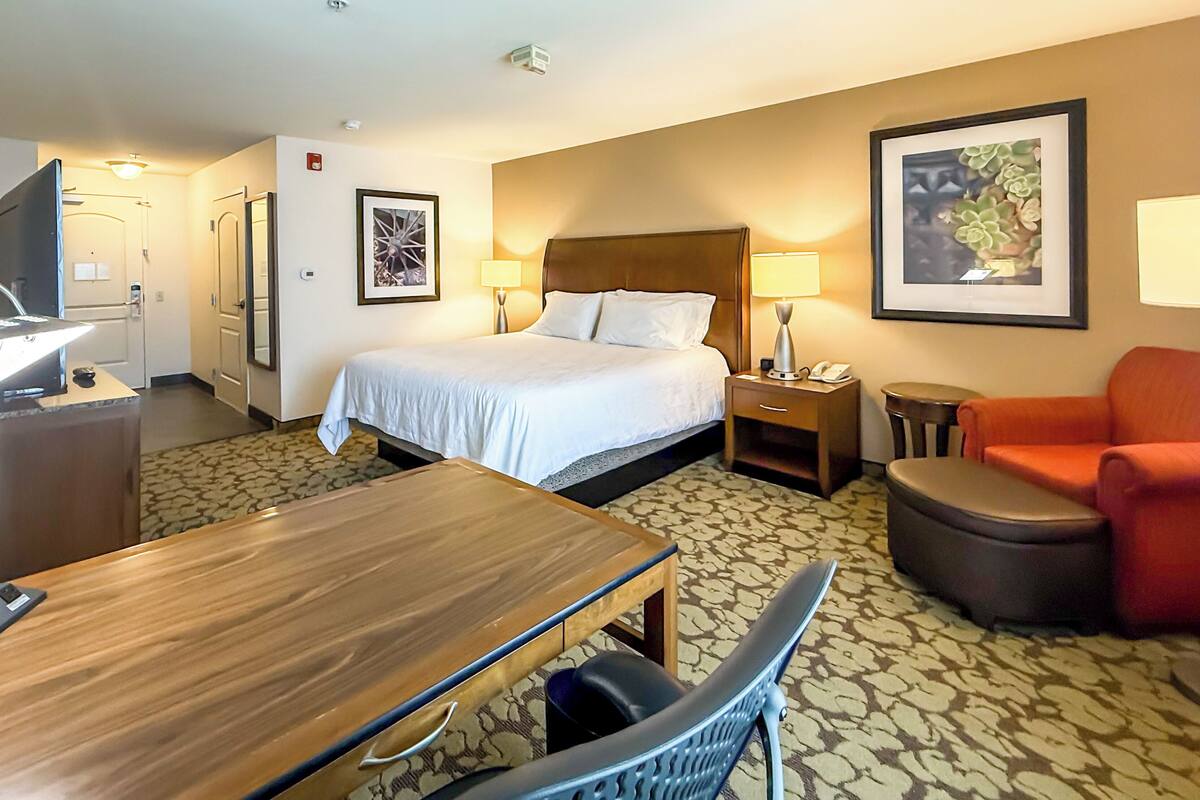 A hotel room with a bed, bedside table, chair, desk, and a small table with a lamp.