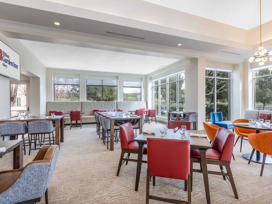 A spacious hotel dining area with multiple tables and chairs, large windows offering views of the outdoors, and a modern ceiling design.