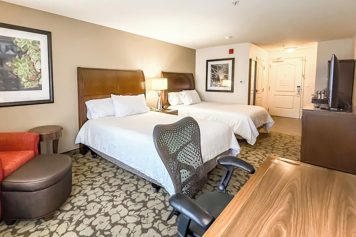 A hotel room with two beds, a desk, a chair, a television, and artwork on the walls.