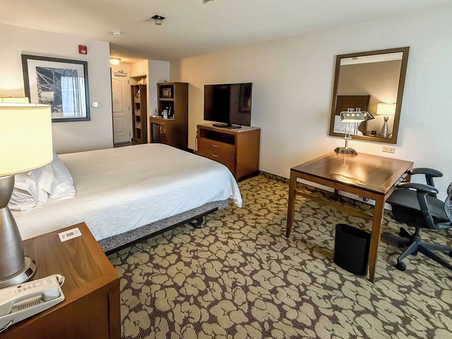 A hotel room with a bed, desk, chair, lamp, TV, and a patterned carpet.