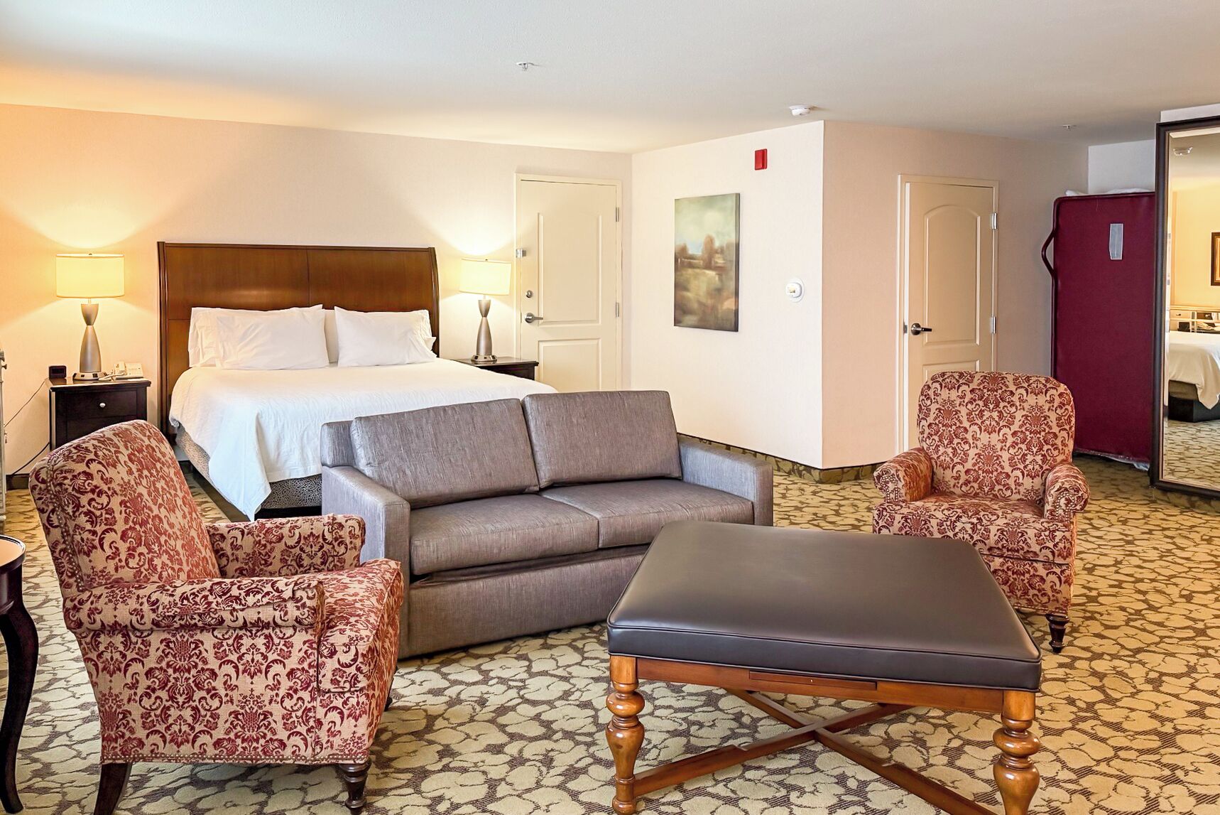 Una habitación de hotel con una cama, dos sillones estampados, un sofá, una mesa de centro de madera y suelo alfombrado.
