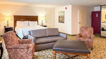 Una habitación de hotel con una cama, dos sillones estampados, un sofá, una mesa de centro de madera y suelo alfombrado.
