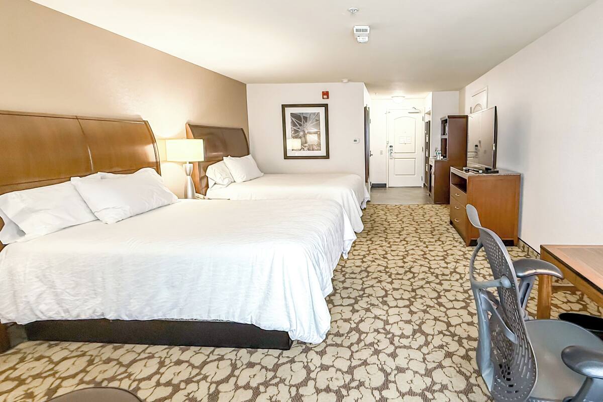 A hotel room with two beds, a desk with a chair, a TV, and a bathroom visible in the background.
