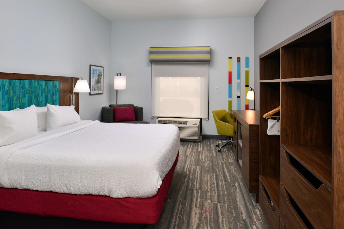 A hotel room with a large bed, a desk, a chair, a wardrobe, and a window with blinds.