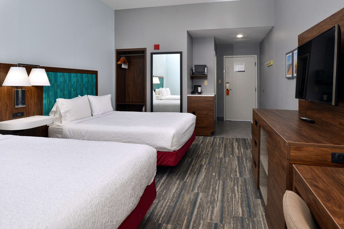 A hotel room with two beds, a desk, a TV, and a bathroom visible through an open door.