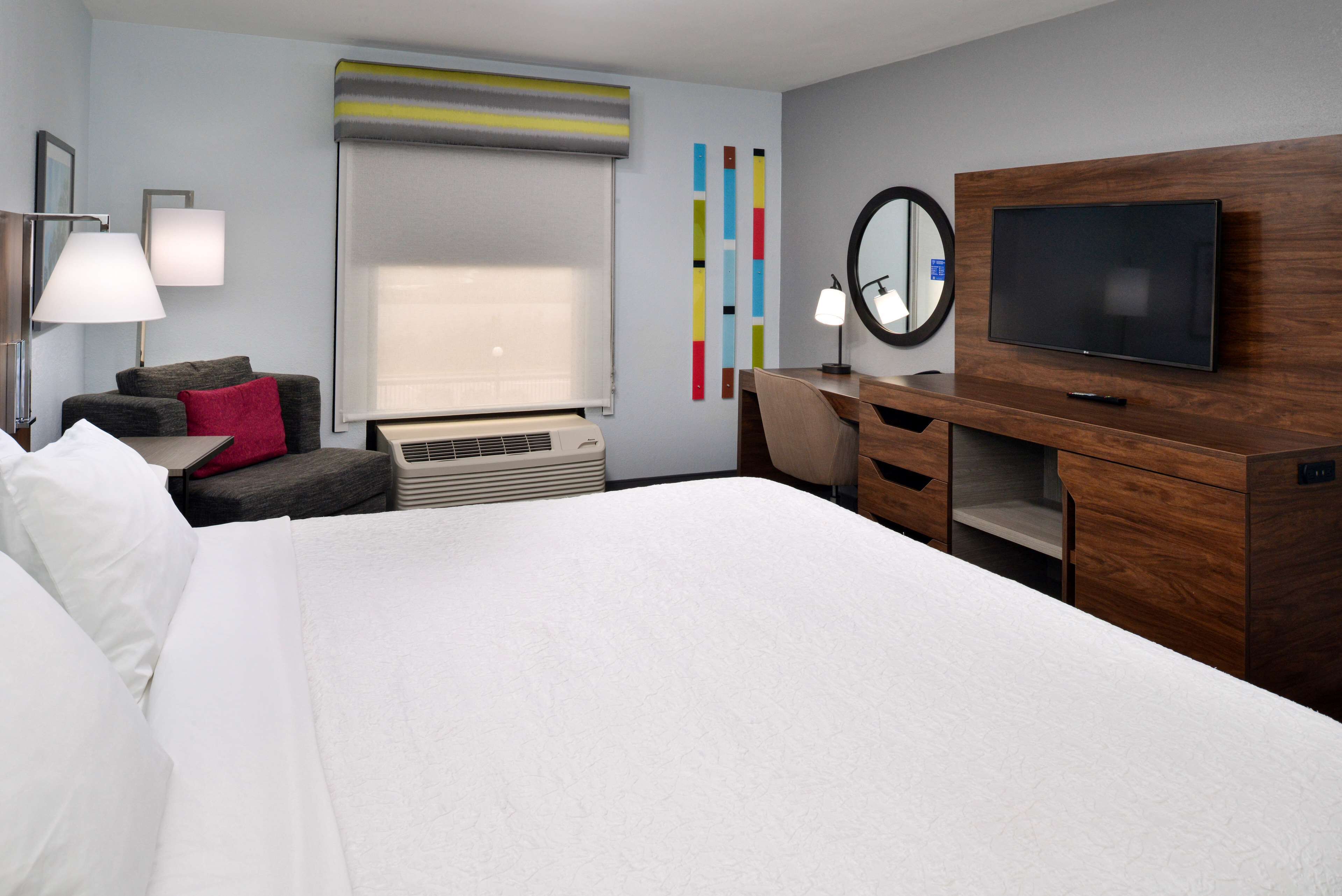 Habitación de hotel con una cama grande, un escritorio con televisor de pantalla plana, una silla, una lámpara, un calentador y una ventana con persianas.