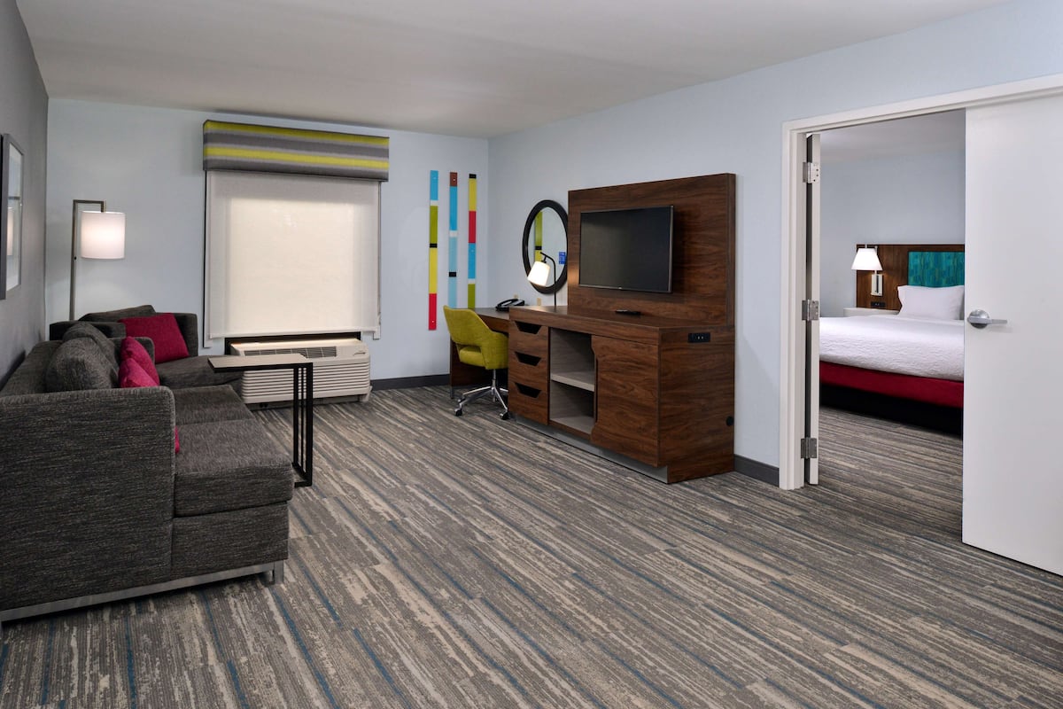 A hotel room with a gray sofa, a wooden desk with a TV, a desk lamp, a chair, a bed, and a window with blinds.