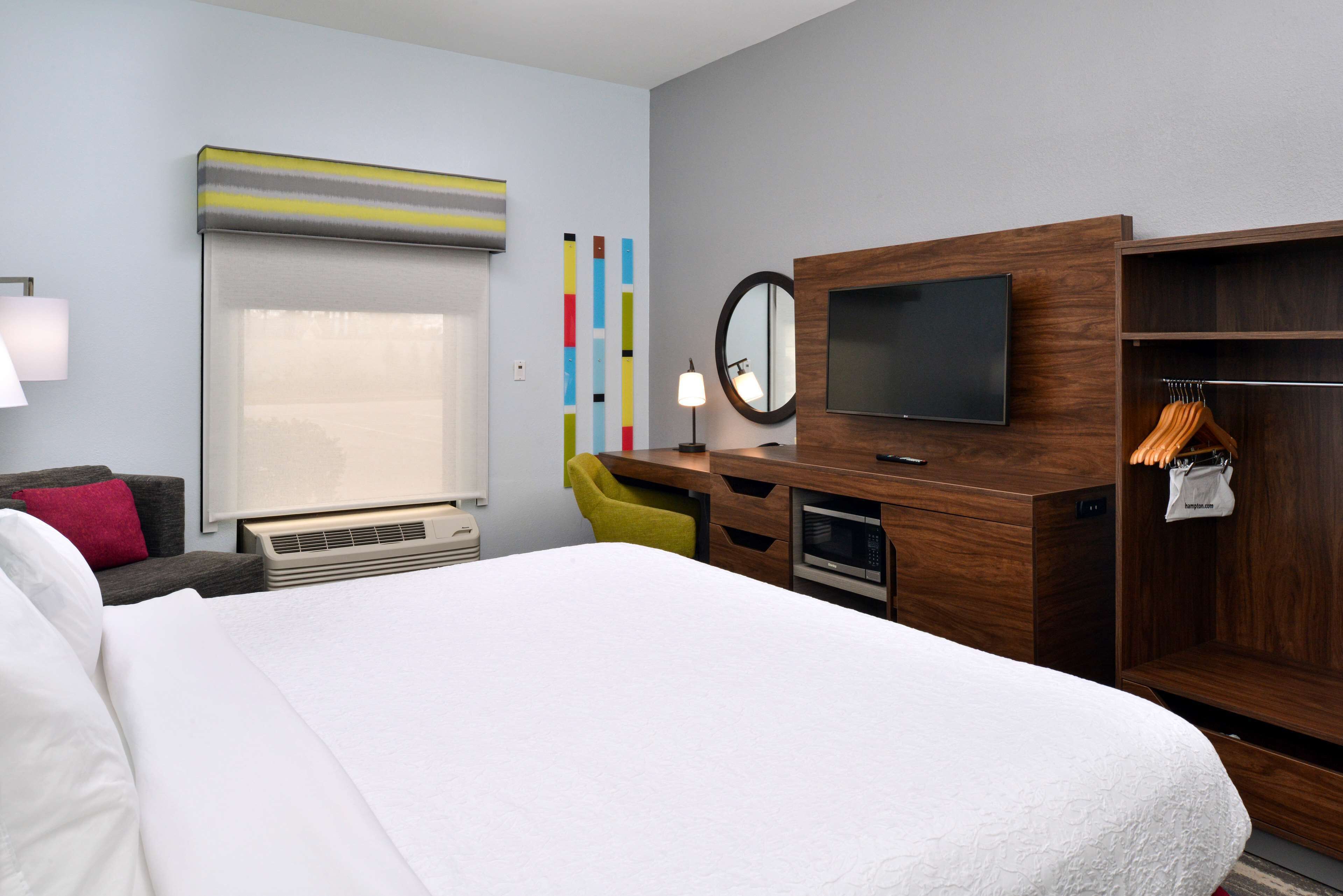 A hotel room with a large bed, a desk with a flat-screen TV, a mirror, a wardrobe, and a seating area with a chair.
