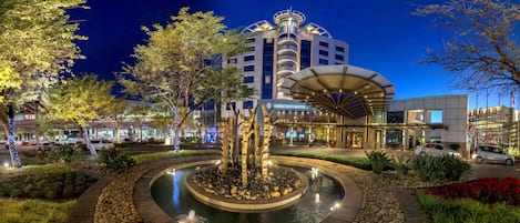 A well-lit outdoor area with a circular water feature, surrounded by landscaped gardens and a modern building with a distinctive architectural design.