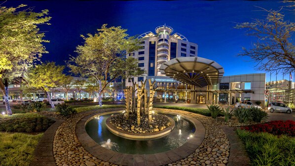 A well-lit outdoor area with a circular water feature, surrounded by landscaped gardens and a modern building with a distinctive architectural design.