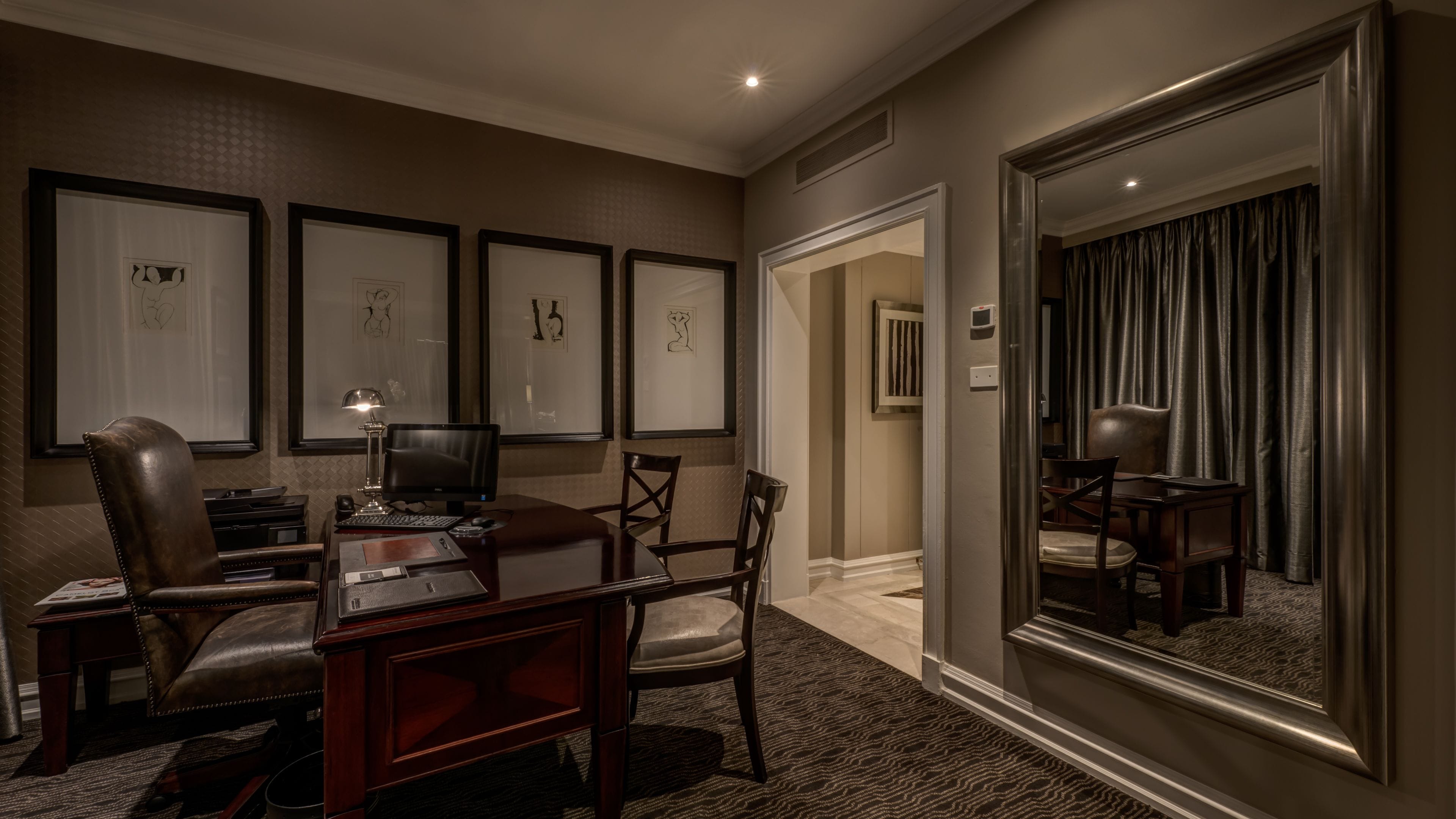 A hotel room with a desk, chair, and a large mirror. There are framed artworks on the wall and a carpeted floor.