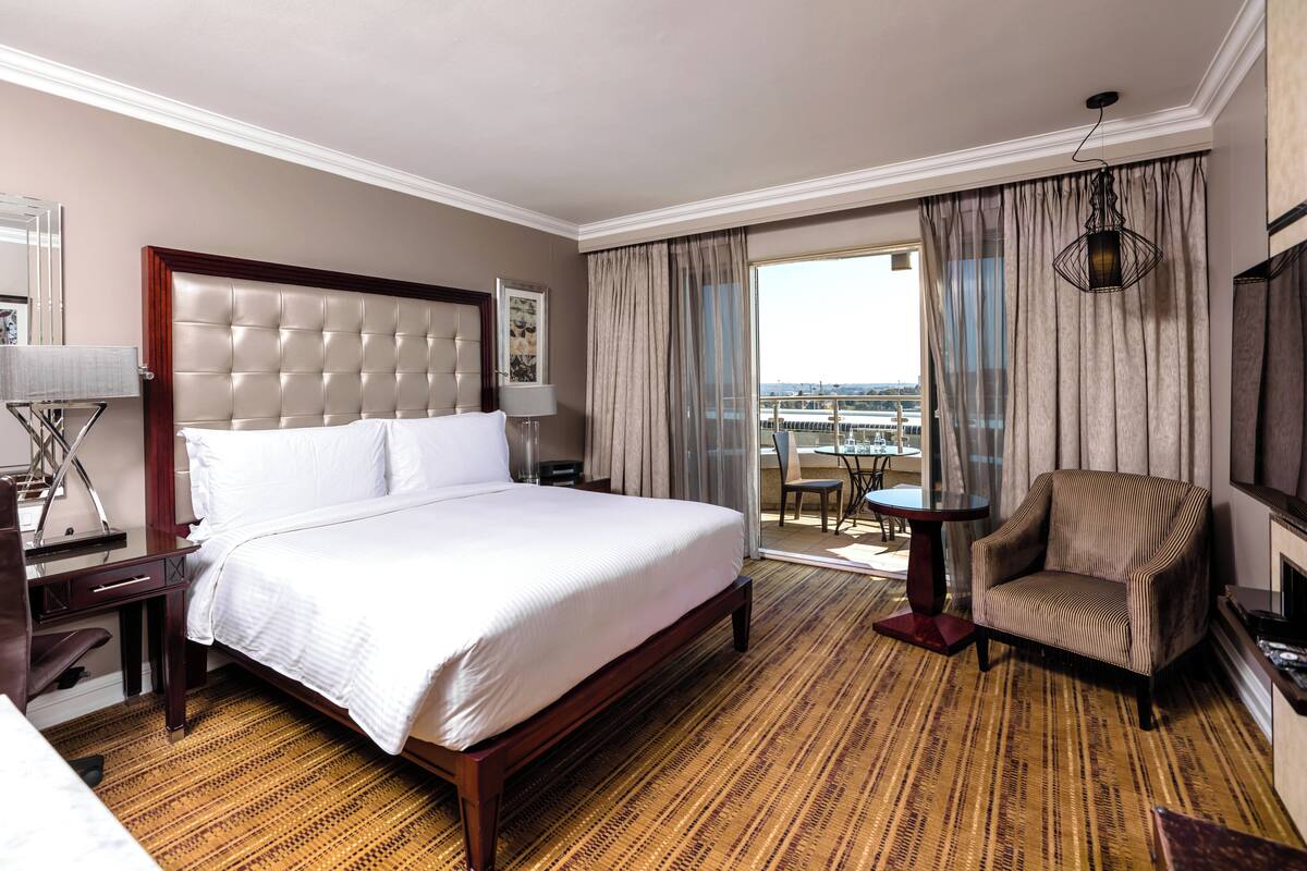 Une chambre d’hôtel avec un grand lit, une chaise, une petite table et une vue sur la ville grâce à de grandes fenêtres.