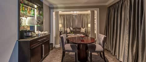 A hotel room with a bar area, a dining table with chairs, a sofa, and a large window with curtains.