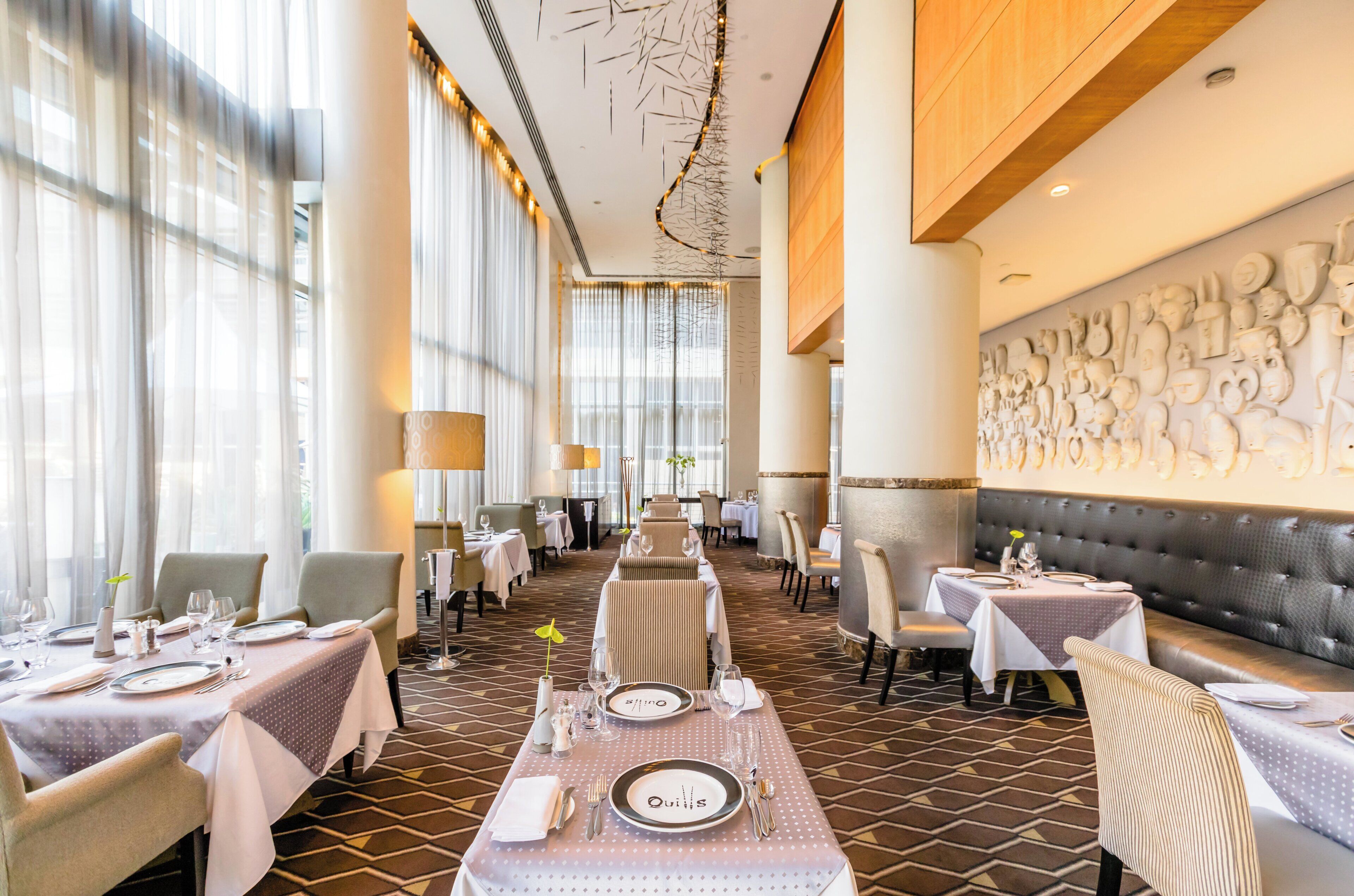 A restaurant with a long table set for dining, multiple seating areas, and a decorative wall with sculptural elements.