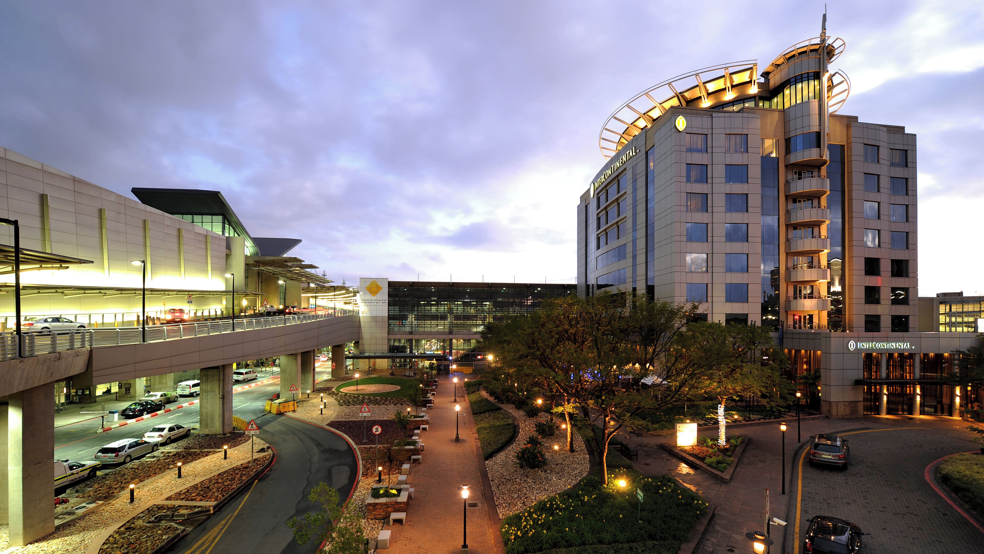 A modern hotel with a covered walkway, landscaped gardens, and a parking area.