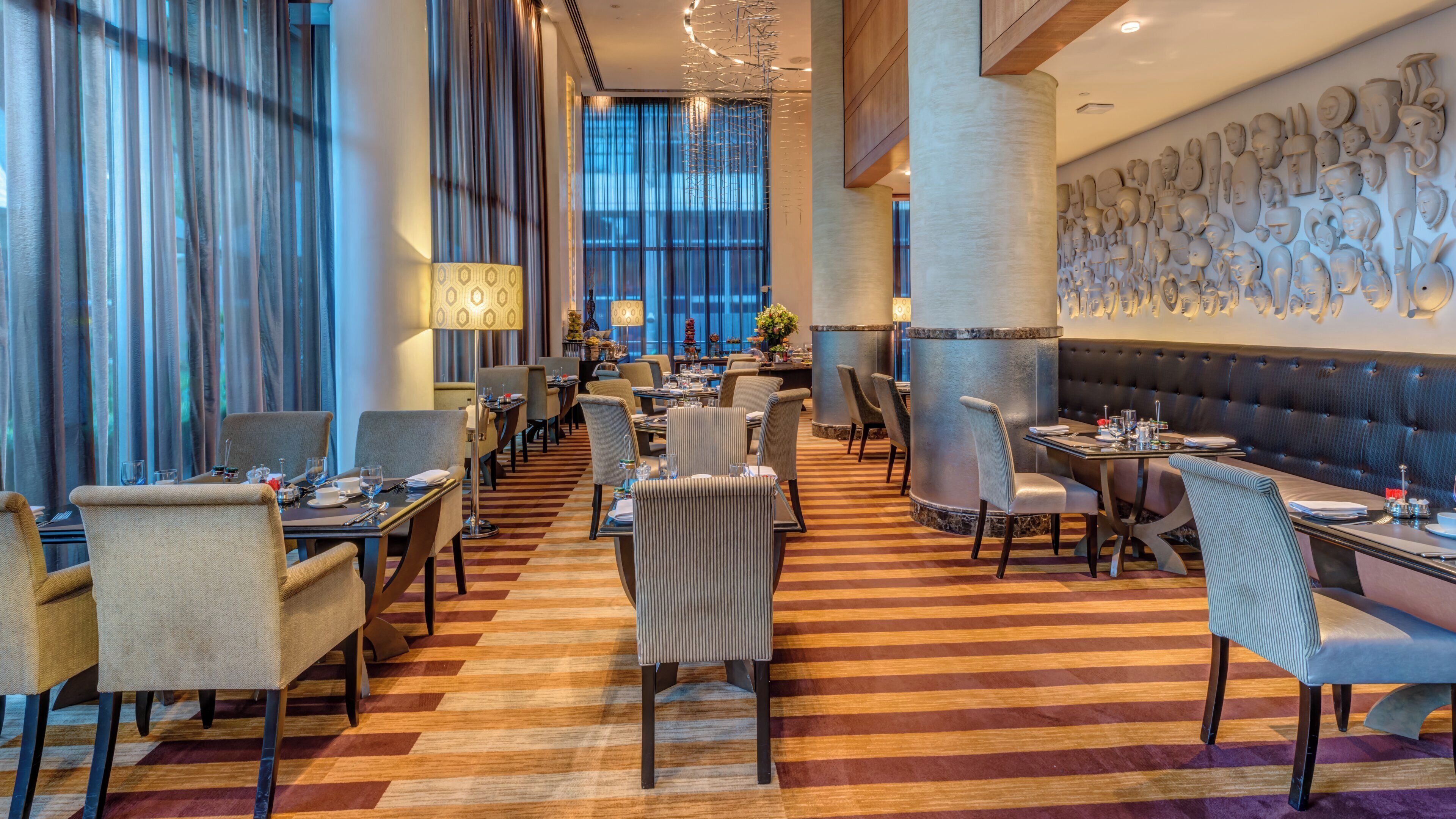 Een restaurant met een lange eetzal, meerdere tafels en stoelen. Er zijn grote ramen met gordijnen, een vloer met een patroon en een wand met sculpturale elementen.