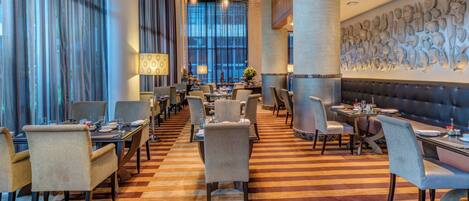 Un restaurante con un amplio salón comedor, múltiples mesas y sillas. Cuenta con ventanas grandes con cortinas, un piso estampado y una pared con elementos decorativos.