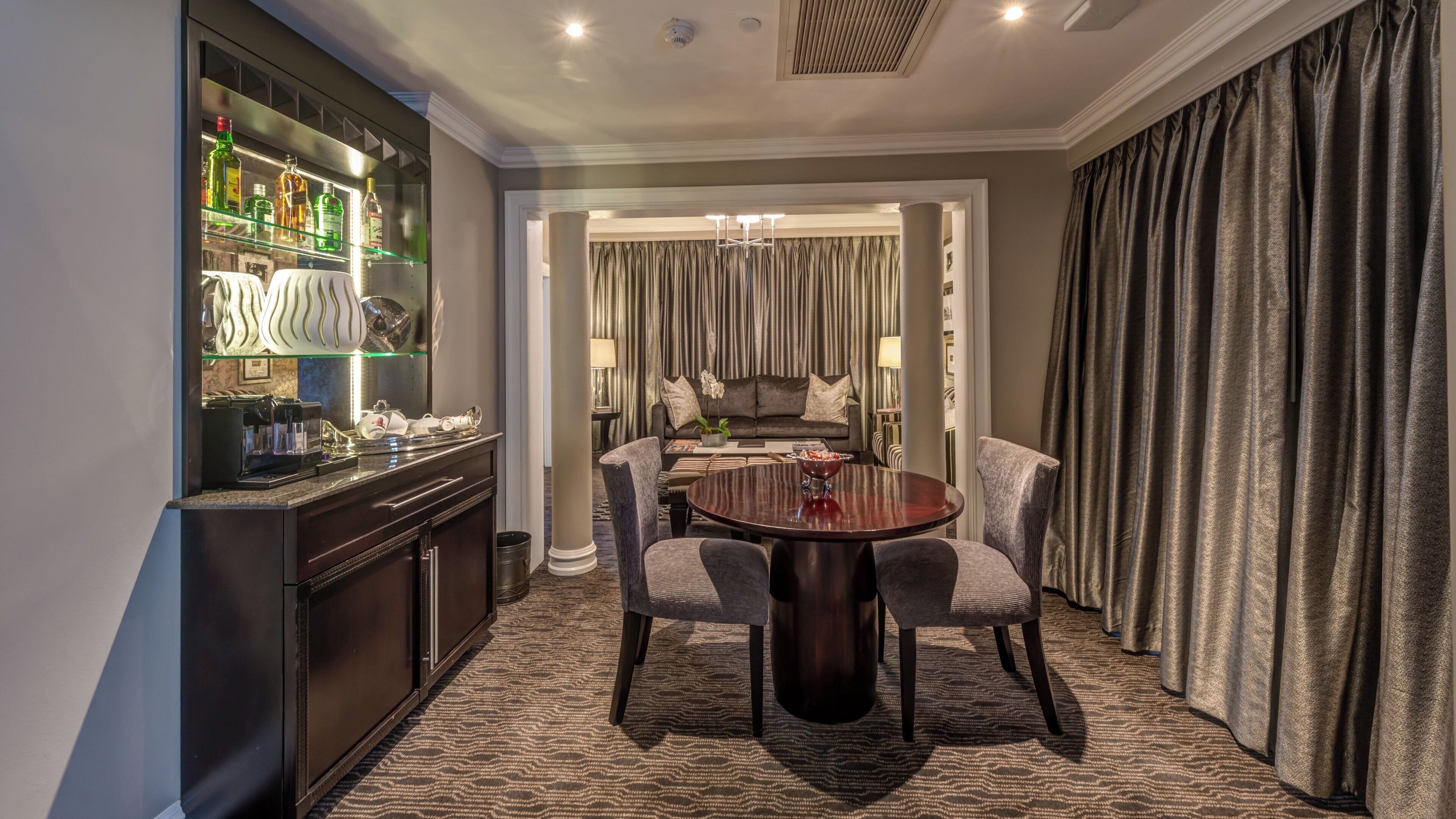 A hotel room with a bar area, a dining table with chairs, a sofa, and a large window with curtains.