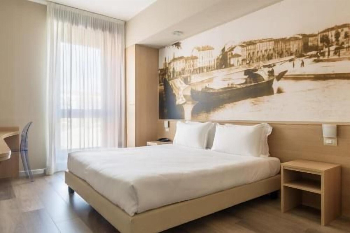 A hotel room with a bed, a nightstand, a chair, a desk, and a large wall mural.