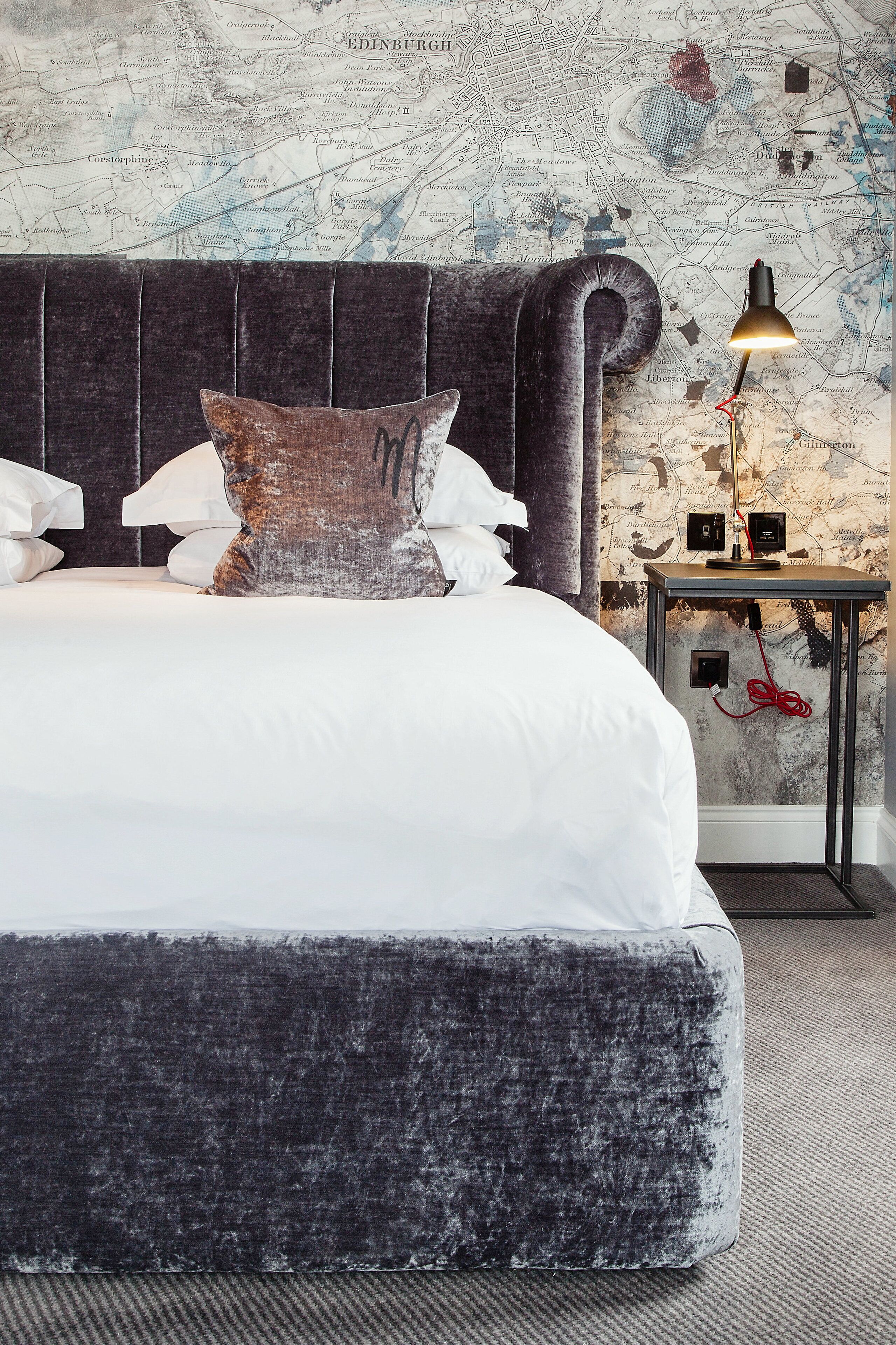 A neatly made bed with a dark headboard, a map-themed wallpaper, and a small bedside table with a lamp.