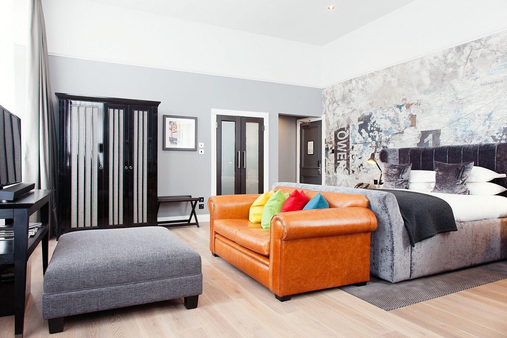 A modern hotel room with a grey sofa, a brown leather couch, a grey armchair, a bed with white and grey bedding, and a large abstract wall art piece.