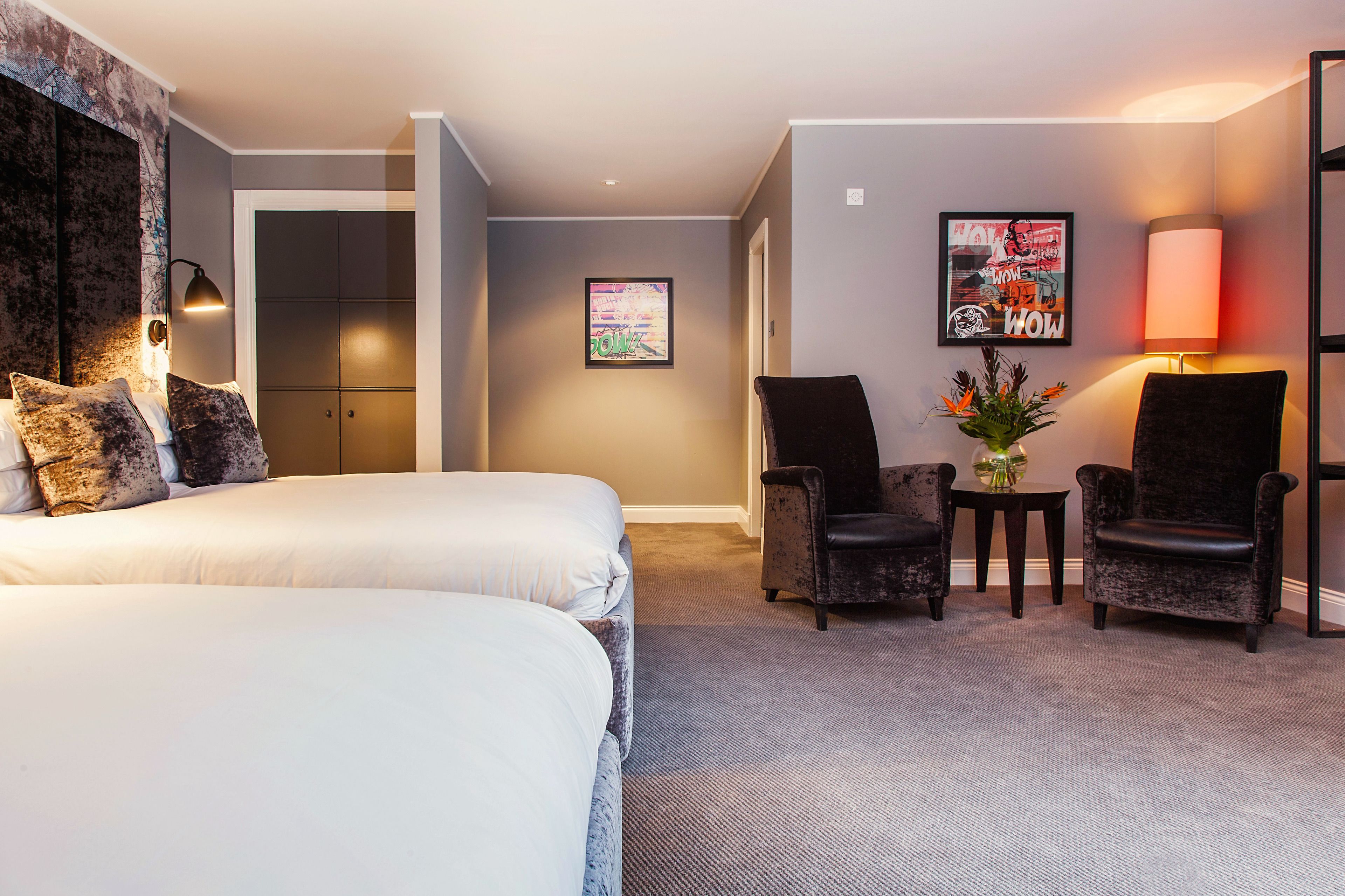 Quarto de hotel com uma cama grande, duas poltronas, uma mesa pequena com um vaso e um abajur.