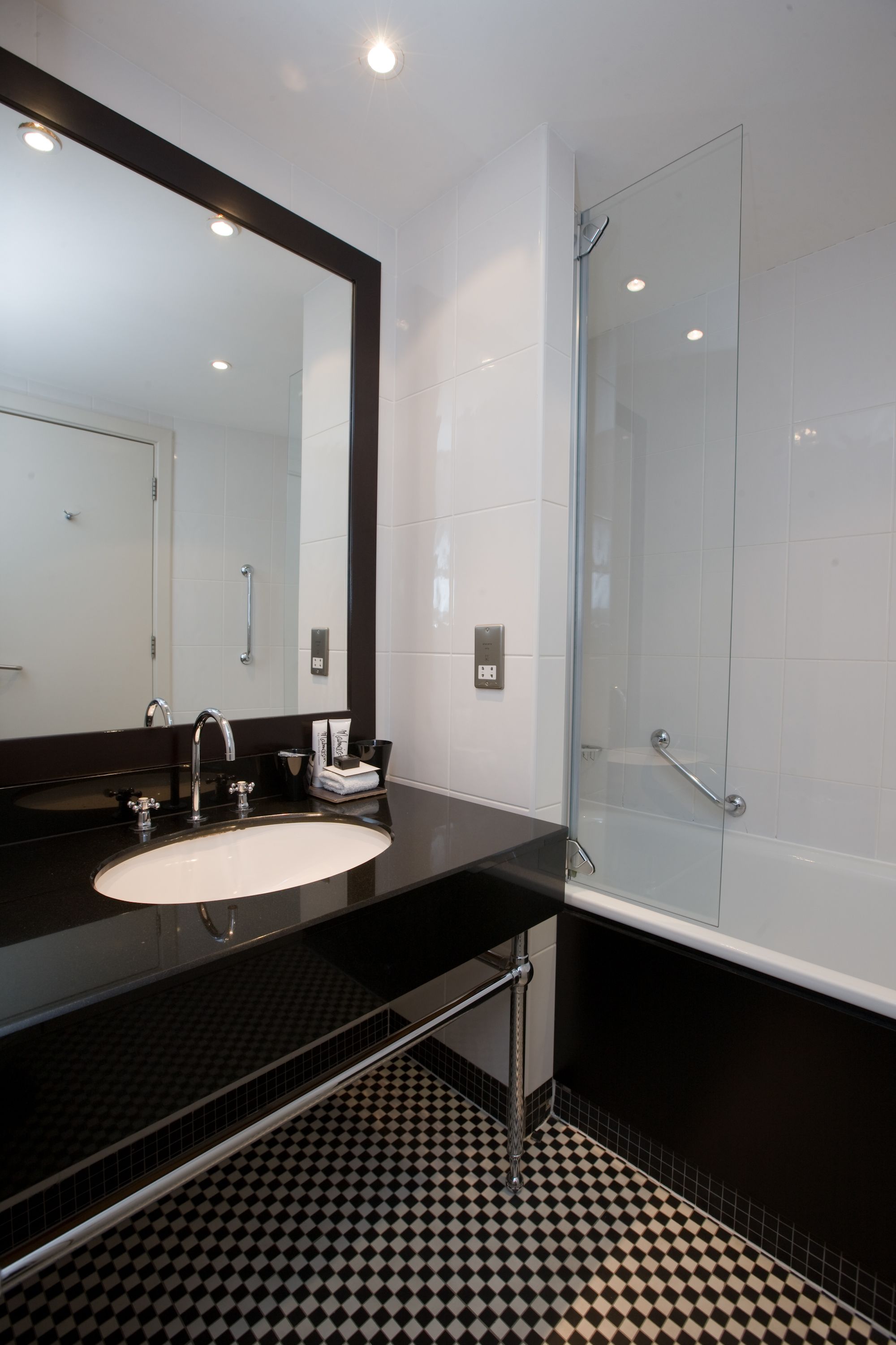 A modern bathroom with a black and white checkered floor, a large mirror, a black countertop, a white sink, and a glass shower enclosure.