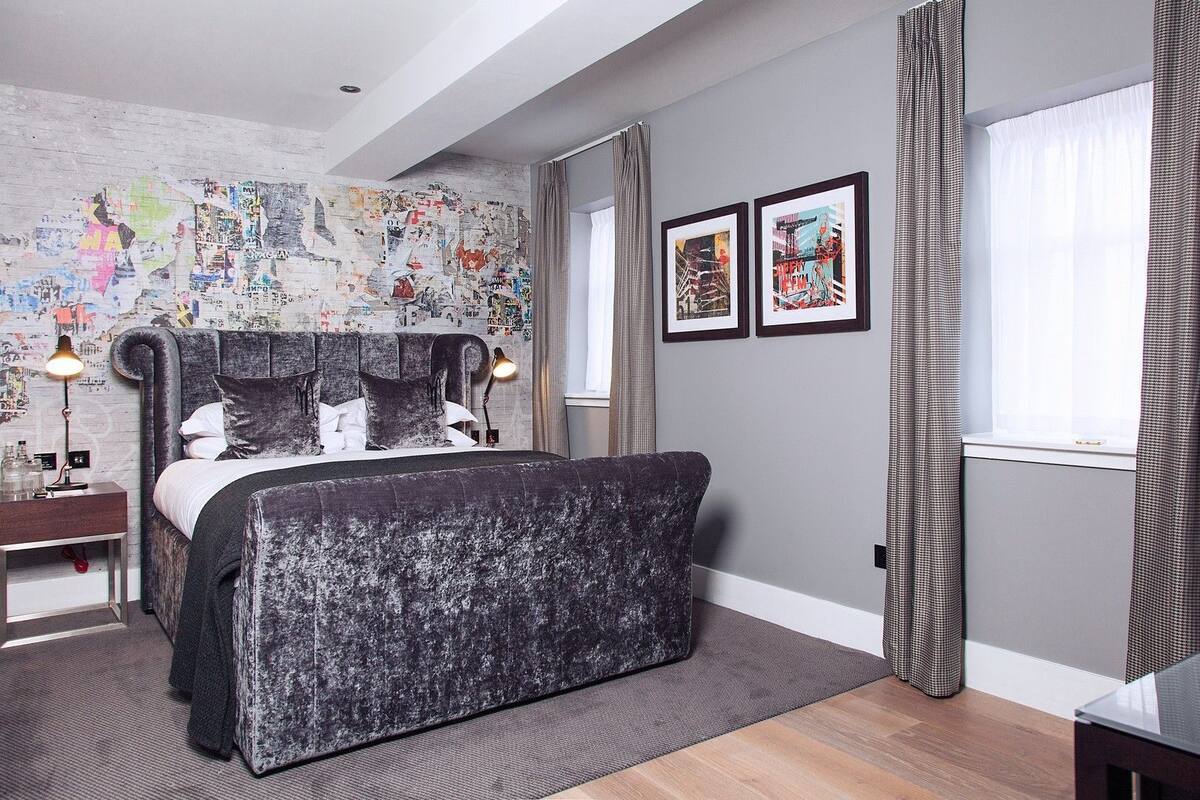 A modern bedroom with a large bed, a headboard with a cityscape mural, a bedside table with a lamp, and a window with curtains.