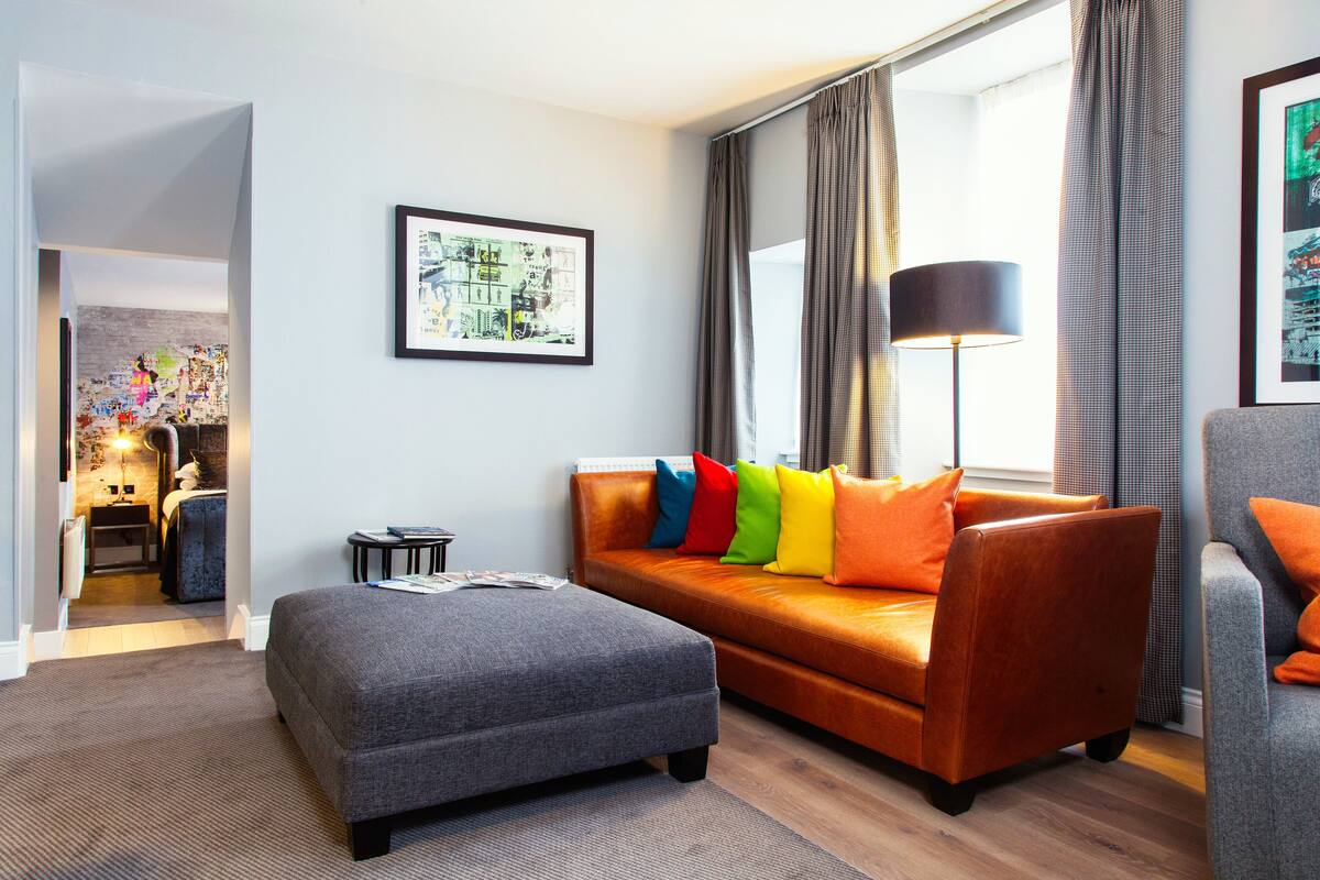 A modern living room with a grey sofa, colorful cushions, a grey ottoman, a black floor lamp, and a large window with curtains.