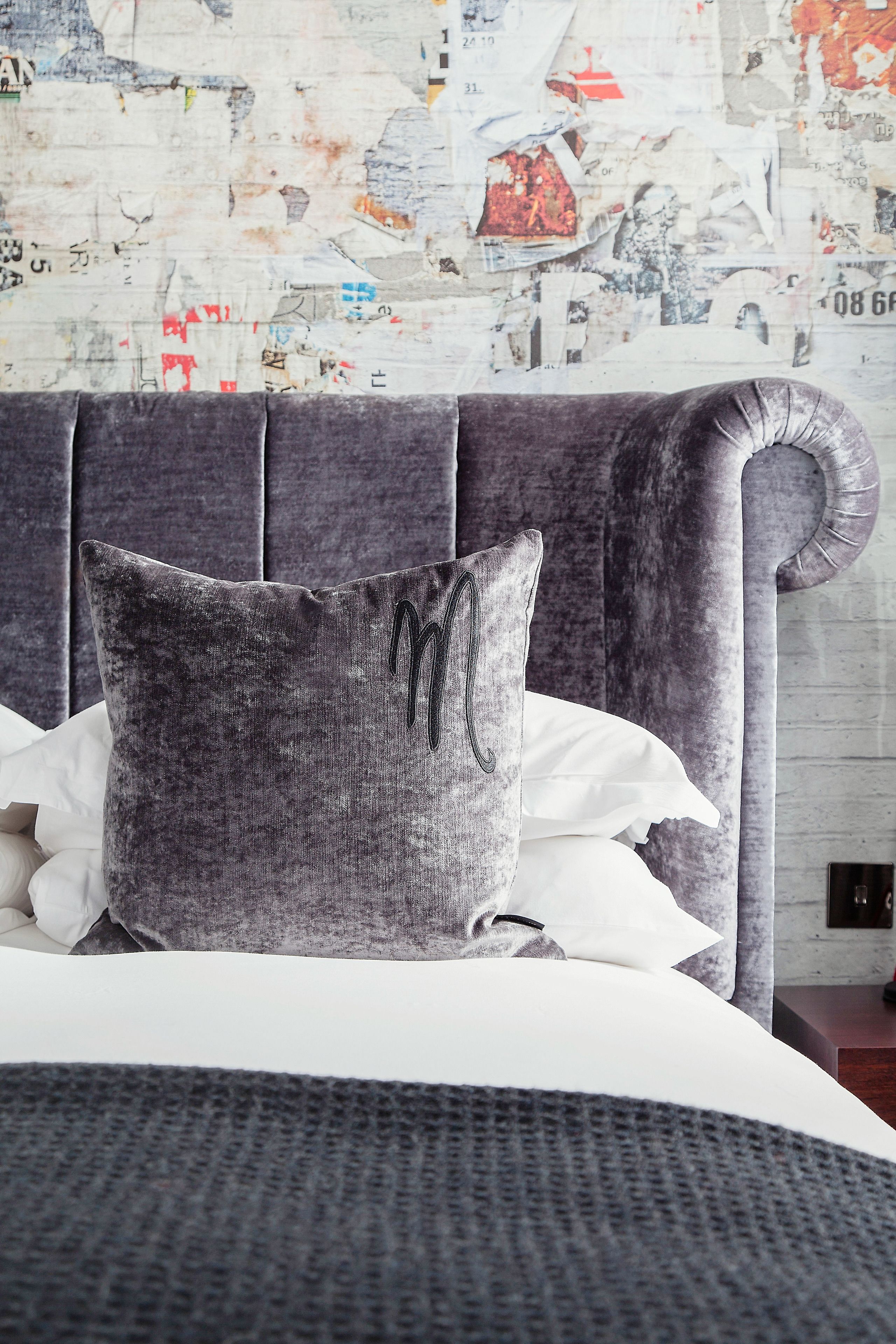 A bed with a grey upholstered headboard, white bedding, and a grey pillow with a monogram.