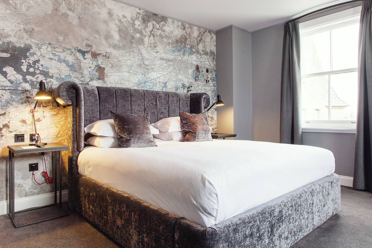 A large bed with a headboard, two bedside lamps, a map on the wall, and a window with curtains.