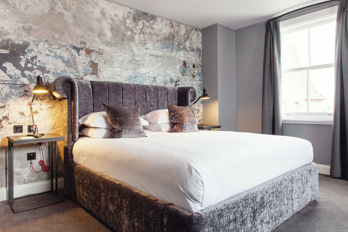 A large bed with a headboard, two bedside tables with lamps, a map on the wall, and a window with curtains.