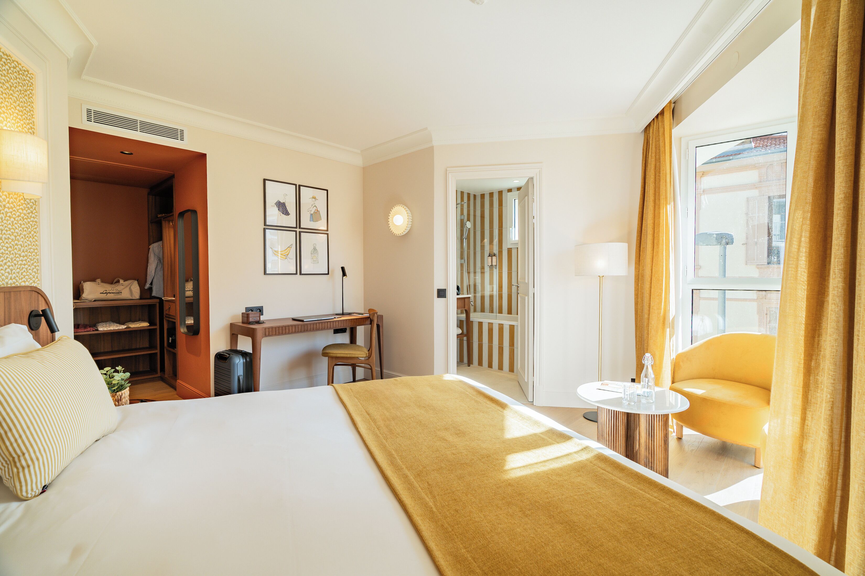 Une chambre d’hôtel comprenant un lit, un bureau, une chaise, une petite table, un miroir et une fenêtre avec des rideaux.