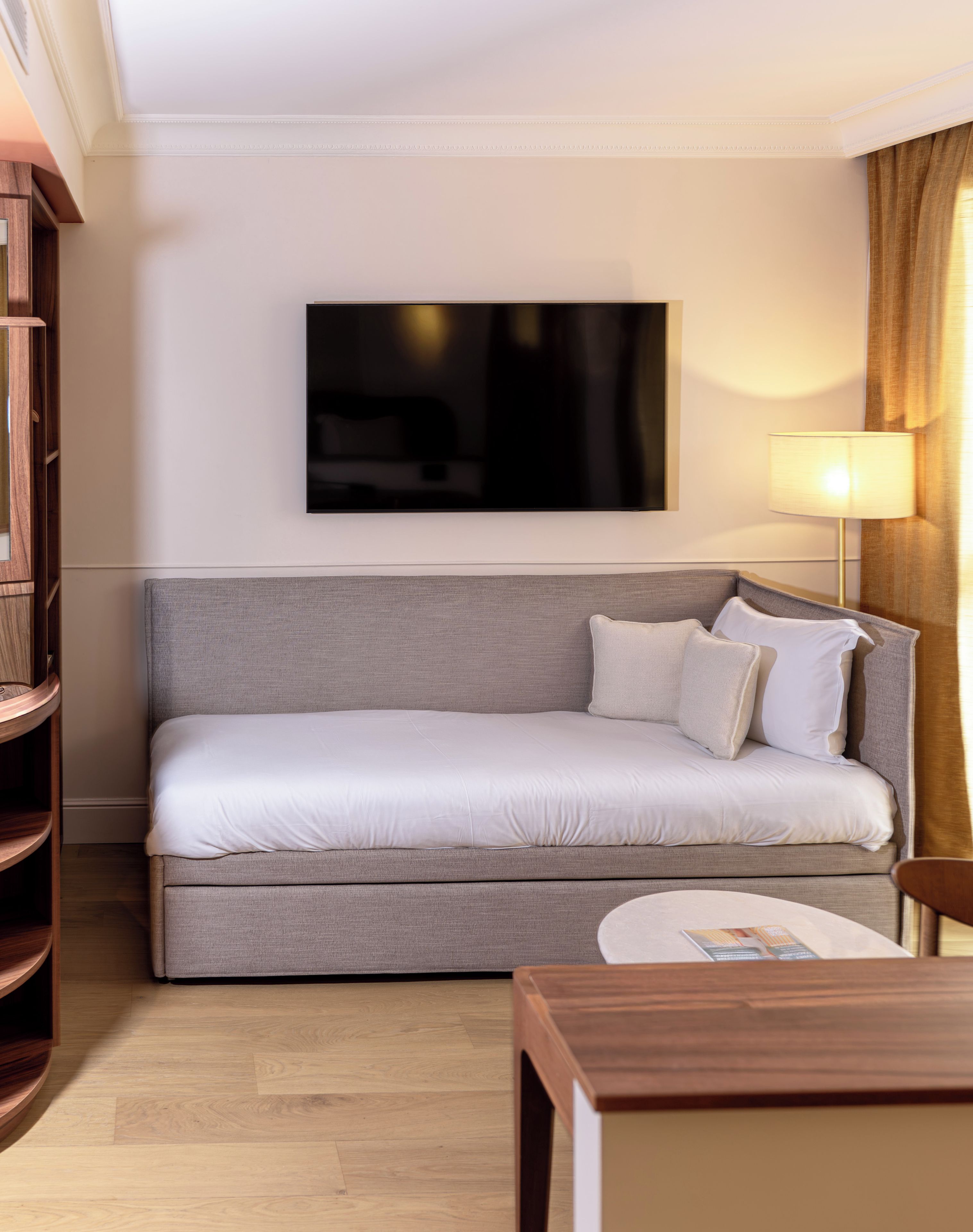 A modern hotel room with a sofa, a flat-screen TV mounted on the wall, a bedside table, and a lamp.