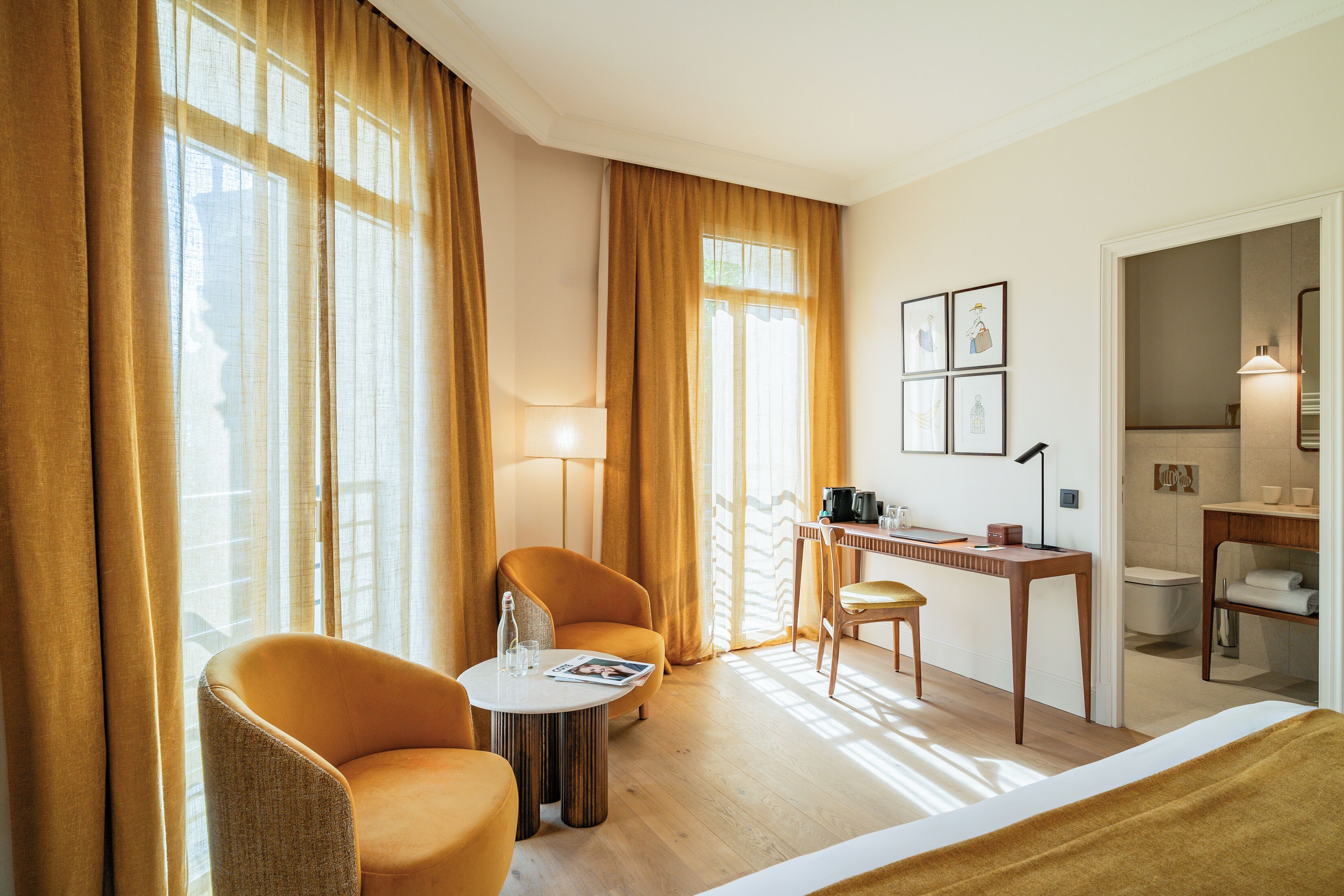 Une chambre d’hôtel avec un lit, deux fauteuils, une petite table ronde, un bureau, une lampe et une salle de bain visible à travers une porte ouverte.