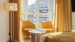 Ein Hotelzimmer mit einem Bett, zwei gelben Sesseln, einem kleinen runden Tisch mit einer Lampe und einem Fenster mit Vorhängen.
