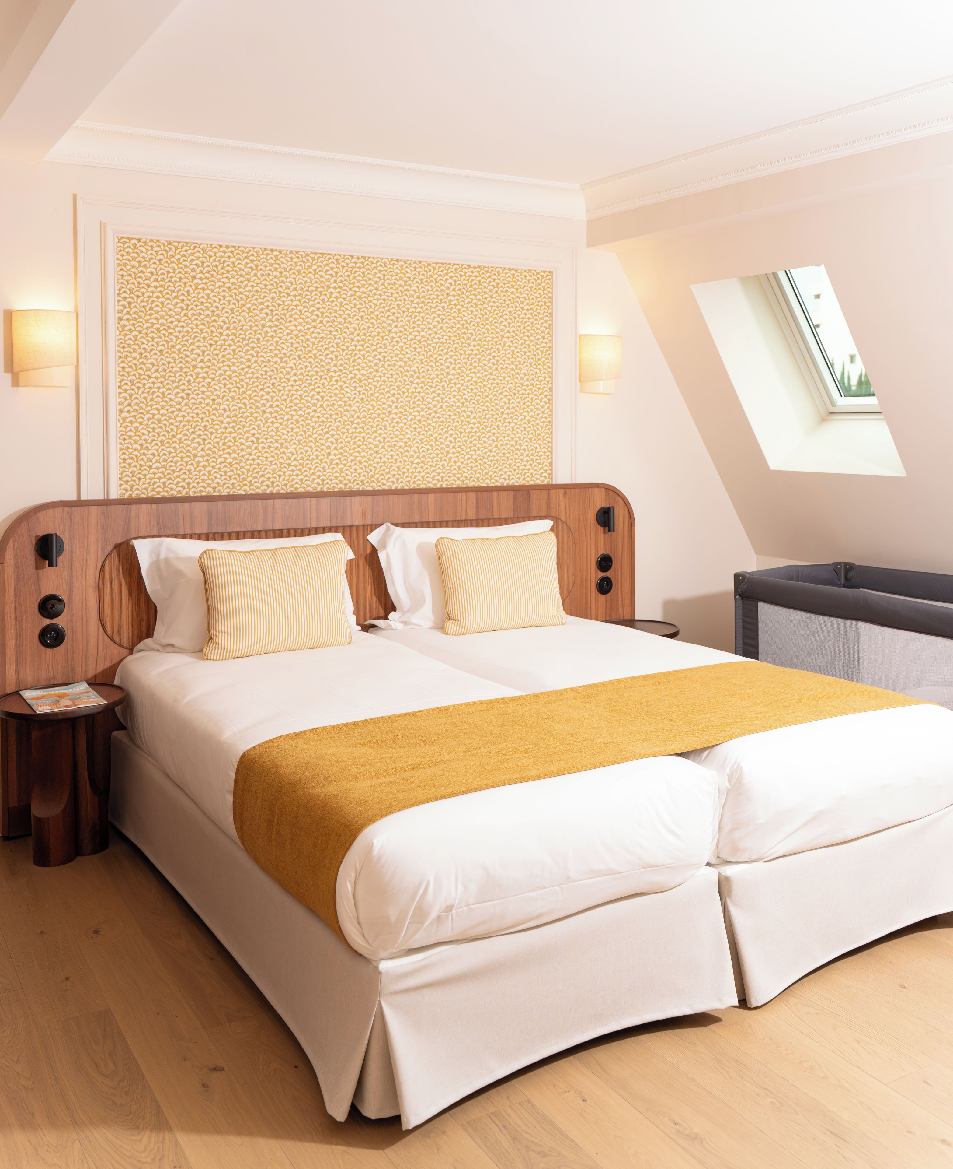 Ein Hotelzimmer mit einem großen Bett, einem hölzernen Kopfteil, zwei Nachttischen, einem kleinen Tischchen, einer Wand mit Muster und einem Oberlicht sowie Blick ins Freie.