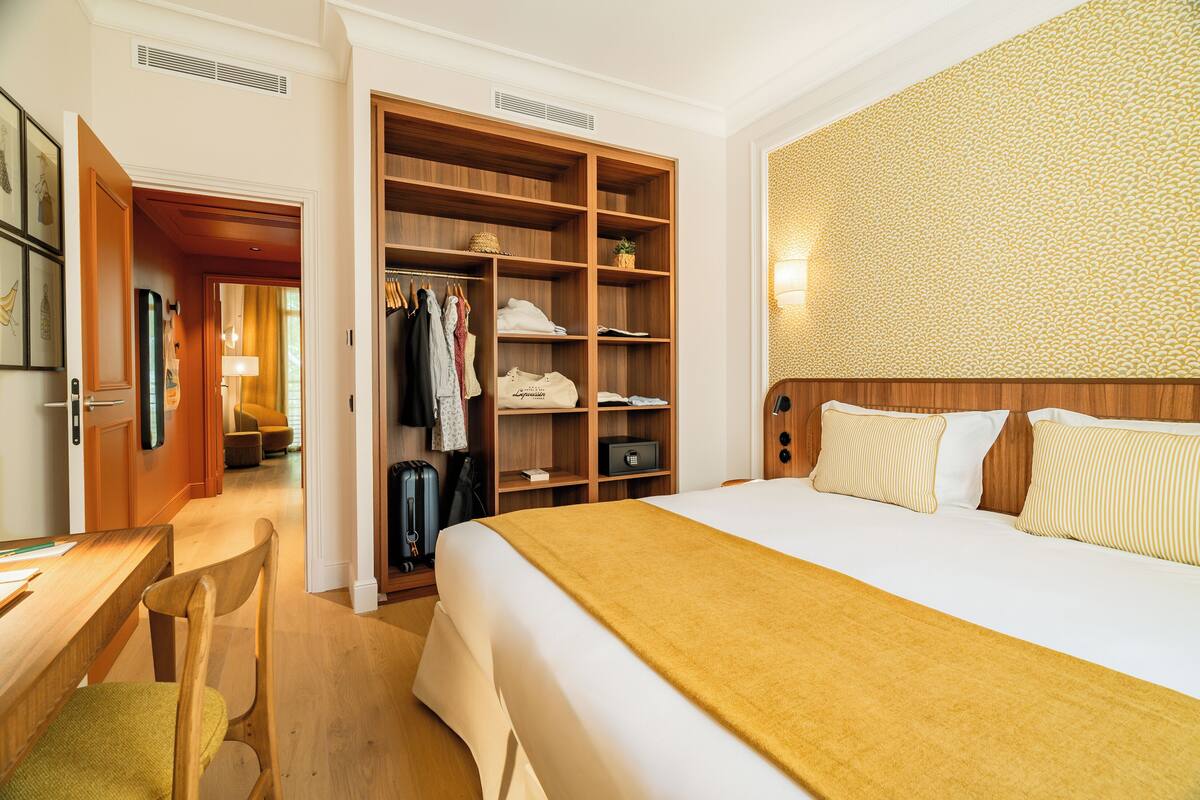 A hotel room with a bed, a wooden wardrobe, a desk with a chair, and a patterned wall.