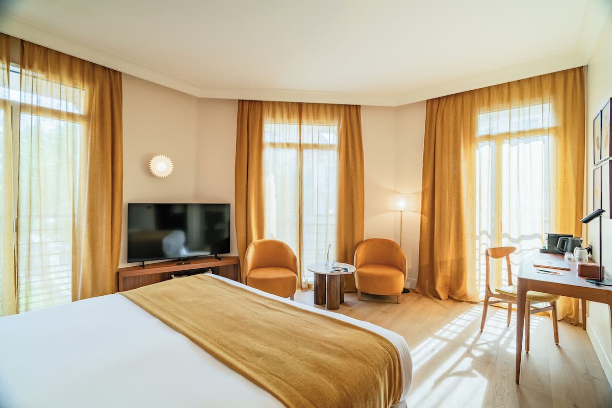 A hotel room with a bed, two armchairs, a desk, a television, and large windows with curtains.