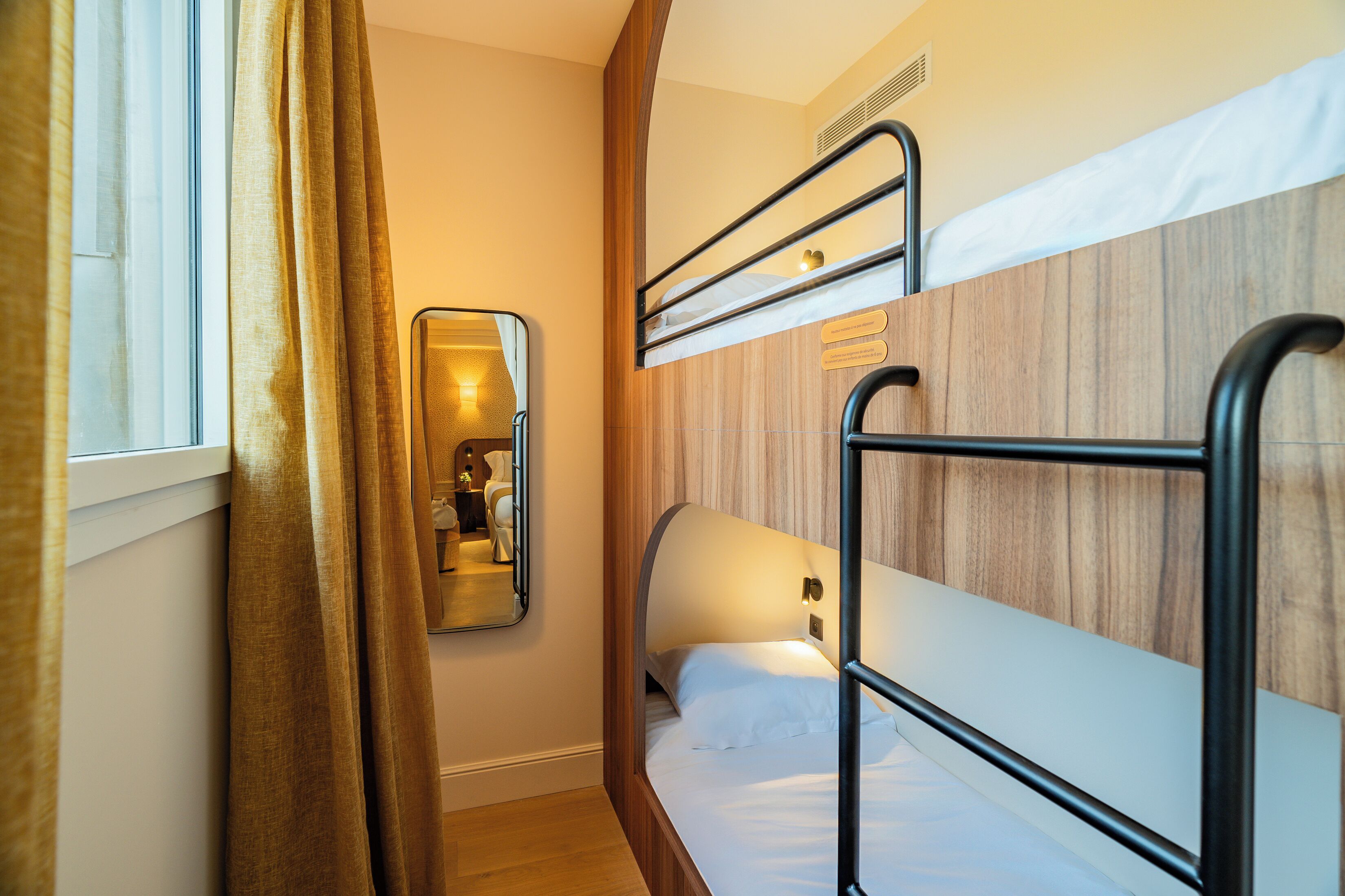 A compact hotel room with bunk beds, a mirror, and a window with curtains.