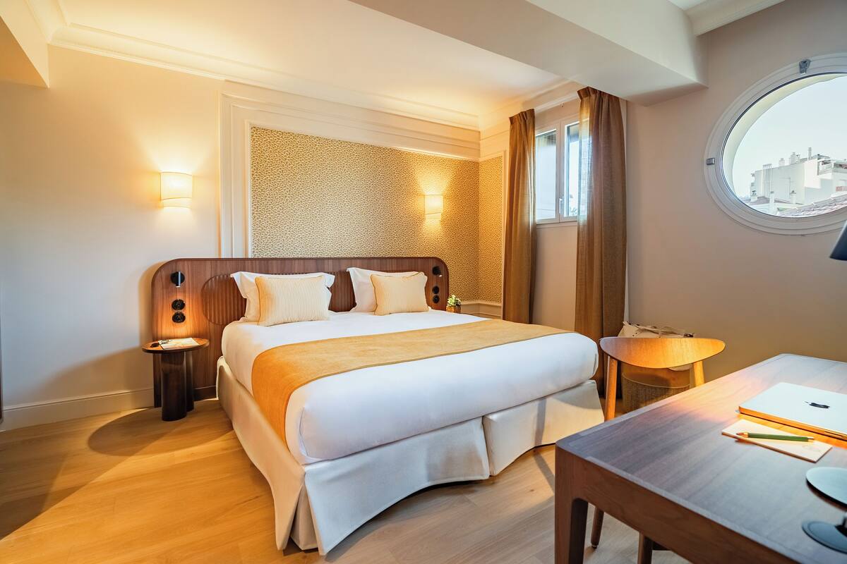 A hotel room with a bed, bedside tables, a desk, a lamp, and a window with a view.