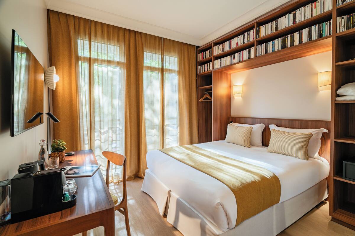 A hotel room with a large bed, a desk with a computer, a chair, a bookshelf, and a large window with curtains.